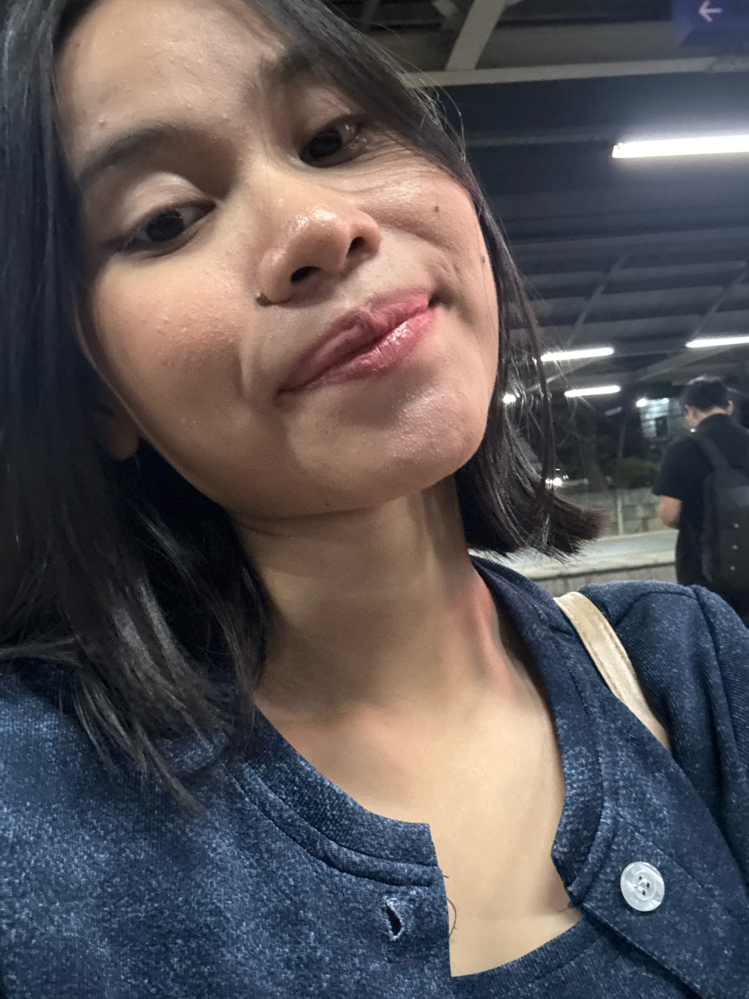 A woman with dark hair and brown eyes is taking a selfie in what appears to be an indoor public space. She is wearing a dark blue sweater. The background is blurred, but it appears to be a station or transit hub with fluorescent lighting and a directional arrow sign. Another person wearing a black shirt and backpack is visible in the background, facing away from the camera. The lighting suggests it is nighttime or a dimly lit indoor environment. The location is identified as East Jakarta, Indonesia.