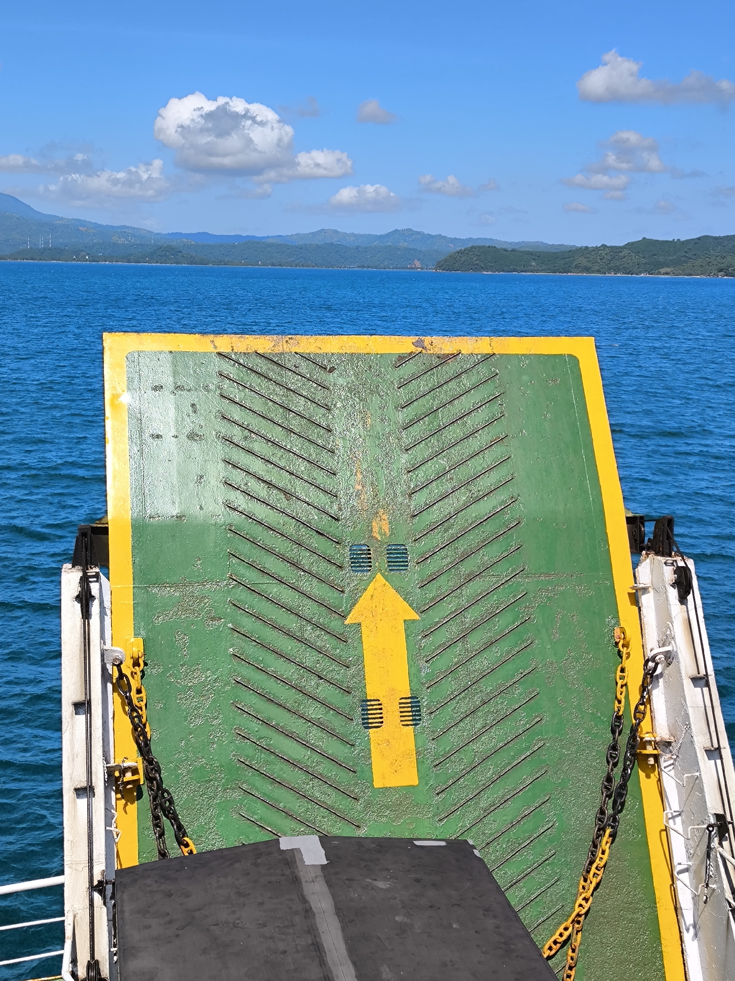 The image shows a ferry ramp extending towards the sea from a boat, with land visible in the distance. The ramp is green with yellow edges and a large yellow arrow pointing upwards. The ramp also has a series of raised black bars for traction. Chains are attached on either side of the ramp. The sea is blue, and the sky is blue with scattered white clouds. The land in the background appears to be hilly and covered with vegetation. This location is in Unknown, Indonesia.