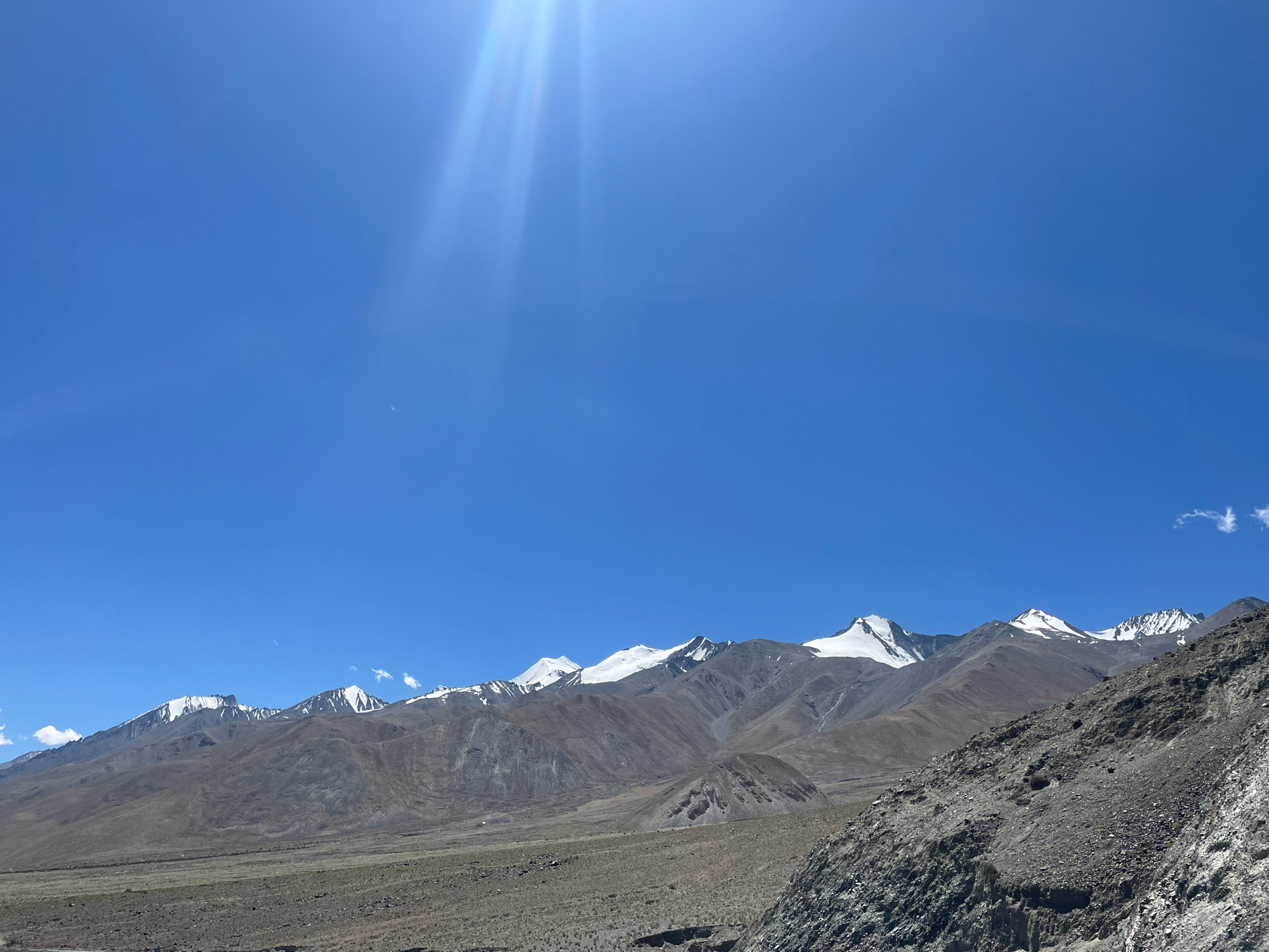 A bright sun shines down on a vast, arid landscape in Spangmik, India. In the foreground, a rocky, gray slope dominates the right side of the frame, with scattered small stones and patches of dry vegetation. Beyond this, a wide, flat expanse of dusty, brown terrain stretches towards a range of imposing mountains. These mountains are characterized by their barren, earthy slopes that transition to snow-capped peaks under the clear, bright blue sky. Sunlight beams streak down from the upper left corner of the image, illuminating the scene with a strong, natural light. No people or animals are visible in the frame.