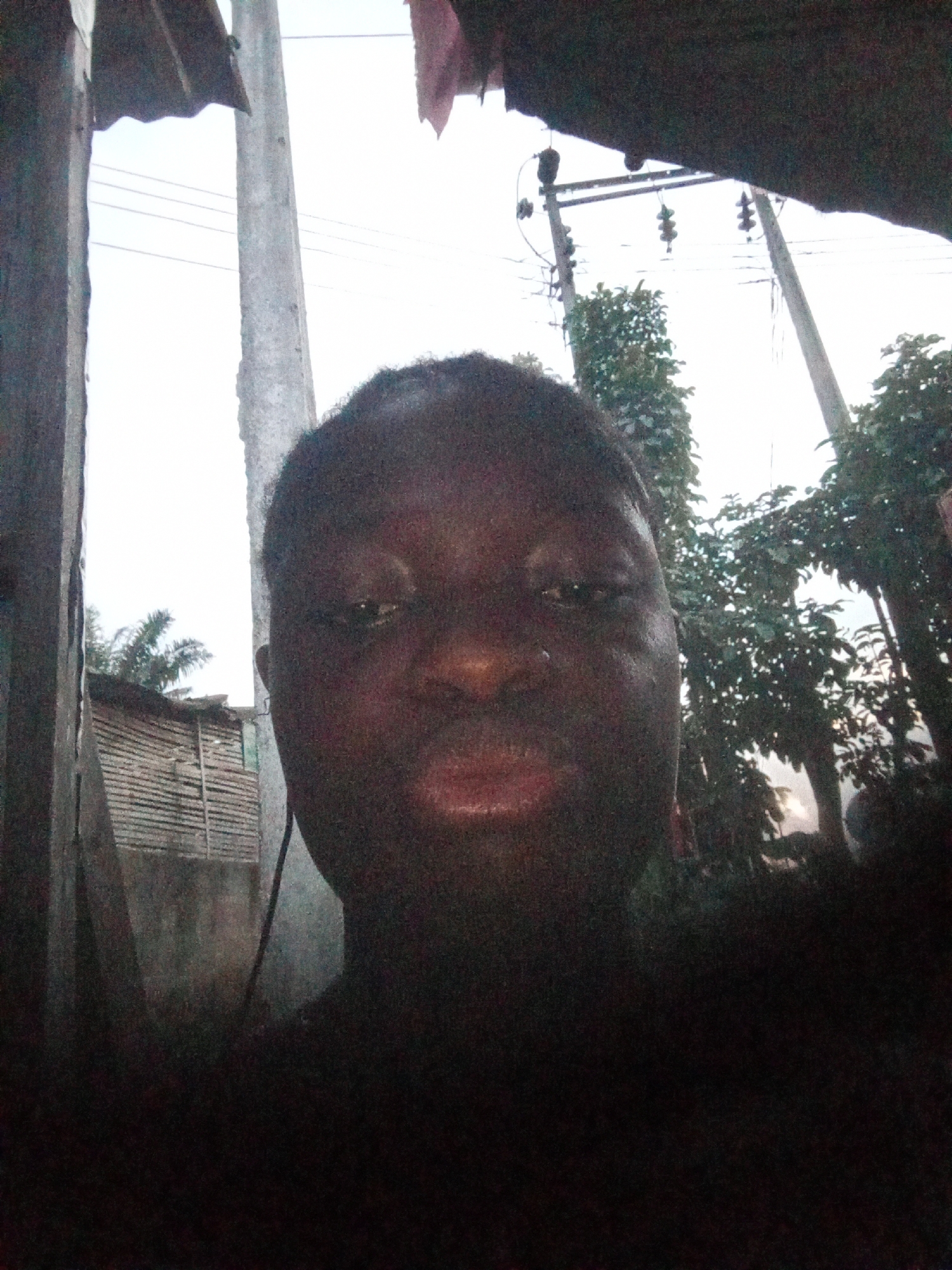 A person with dark skin is taking a selfie in what appears to be a residential area in Mbribit, Nigeria. Overhead power lines and poles are visible. Some vegetation is visible and there is a building made of wood.