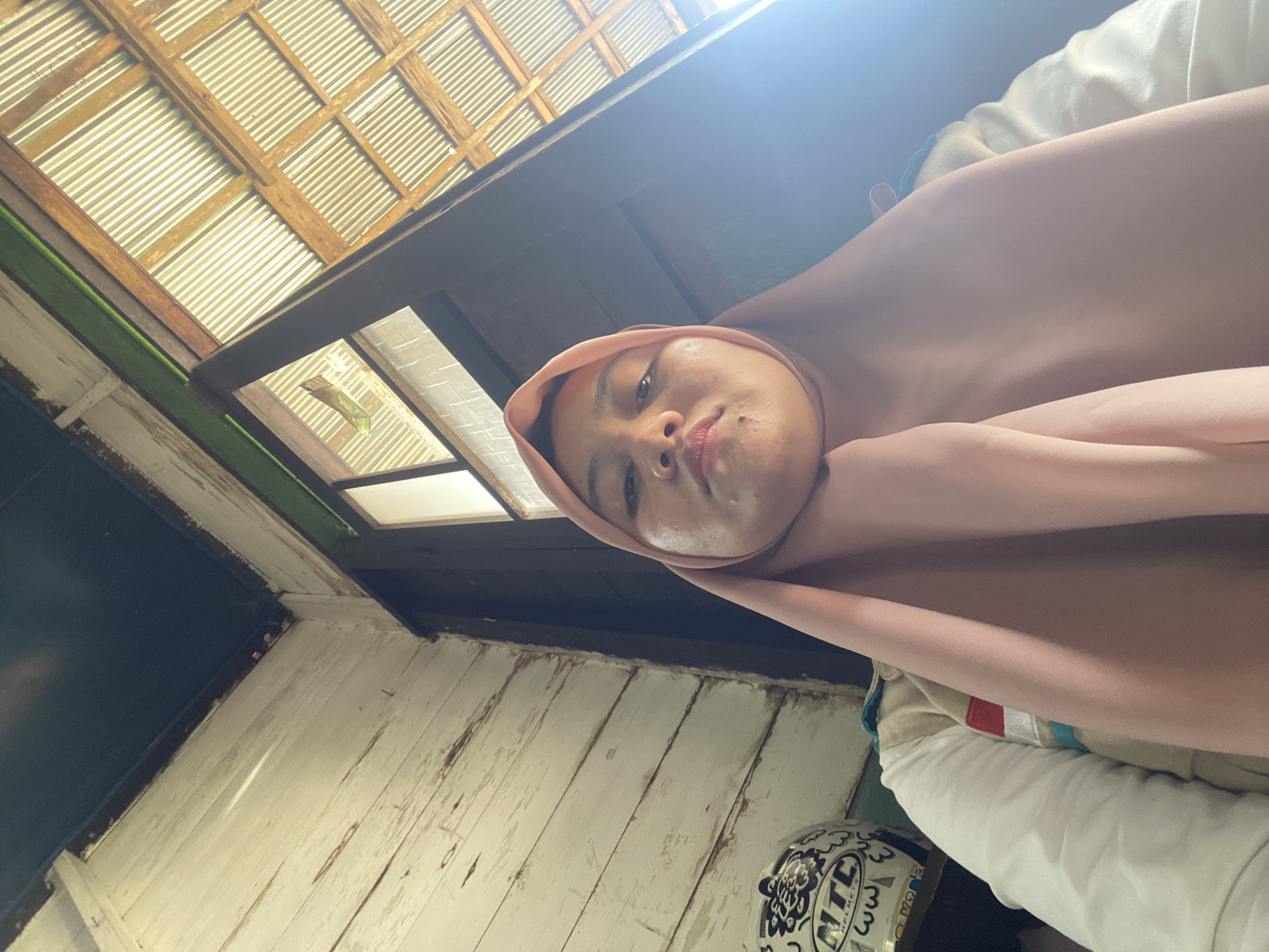 The image is a low-angle, close-up selfie of a young woman wearing a pink hijab and a white shirt with red and blue accents. She is looking directly at the camera with a neutral expression. To her left, partially visible, is a white motorcycle helmet with black and blue designs, including the text "NTC HELMETS". The background consists of rustic wooden walls and a corrugated metal roof, suggesting an outdoor or semi-outdoor setting, possibly a building with an open or partially constructed roof. The lighting is bright, with a lens flare visible on the right side of the image, indicating sunlight. The overall impression is casual and personal. While the prompt mentions Bawahan Selan, Indonesia, there are no specific visual cues within the image to confirm this location.