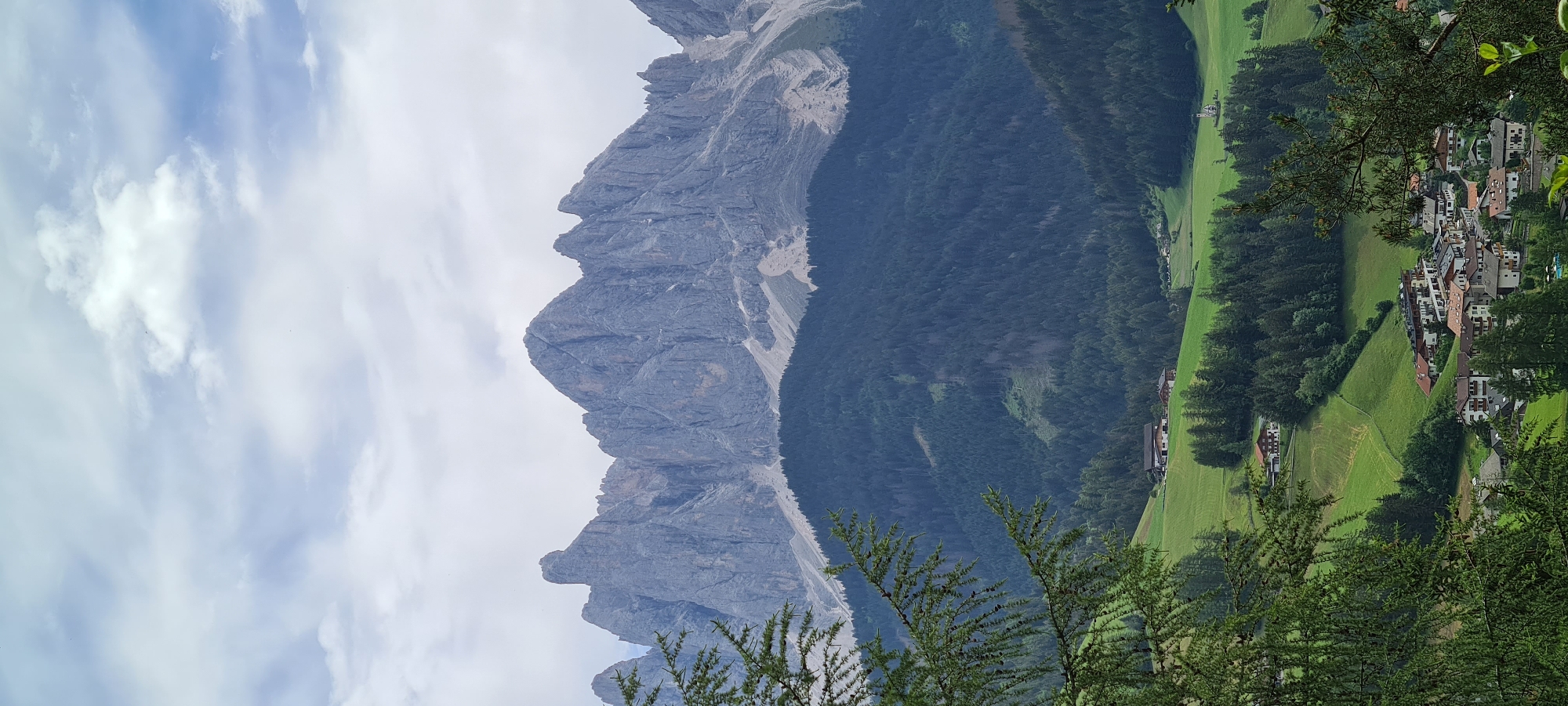 The image shows a panoramic view of the village of St. Magdalena, Santa Maddalena, Italy, nestled in a valley with the dramatic peaks of the Dolomites mountain range in the background. The foreground features the lush green slopes of the valley, dotted with trees and scattered houses. A cluster of buildings, characteristic of the village, is visible in the lower portion of the image, with red-tiled roofs and a mix of traditional and modern architecture.
The middle ground is dominated by a dense forest of dark green pine trees covering the mountainside. Further back, the imposing grey, jagged peaks of the Dolomites rise sharply against a cloudy sky. The sky itself is a mix of white and grey clouds, with patches of blue peeking through, suggesting a partly cloudy day.
The scene is tranquil and picturesque, capturing the natural beauty of the Italian Alps and the charm of a small mountain village. There are no people or animals visible in the frame. The focus is on the landscape, showcasing the geological formations of the Dolomites and the verdant scenery of the valley. The composition draws the viewer's eye from the foreground foliage, across the village and forest, up to the impressive mountain range.