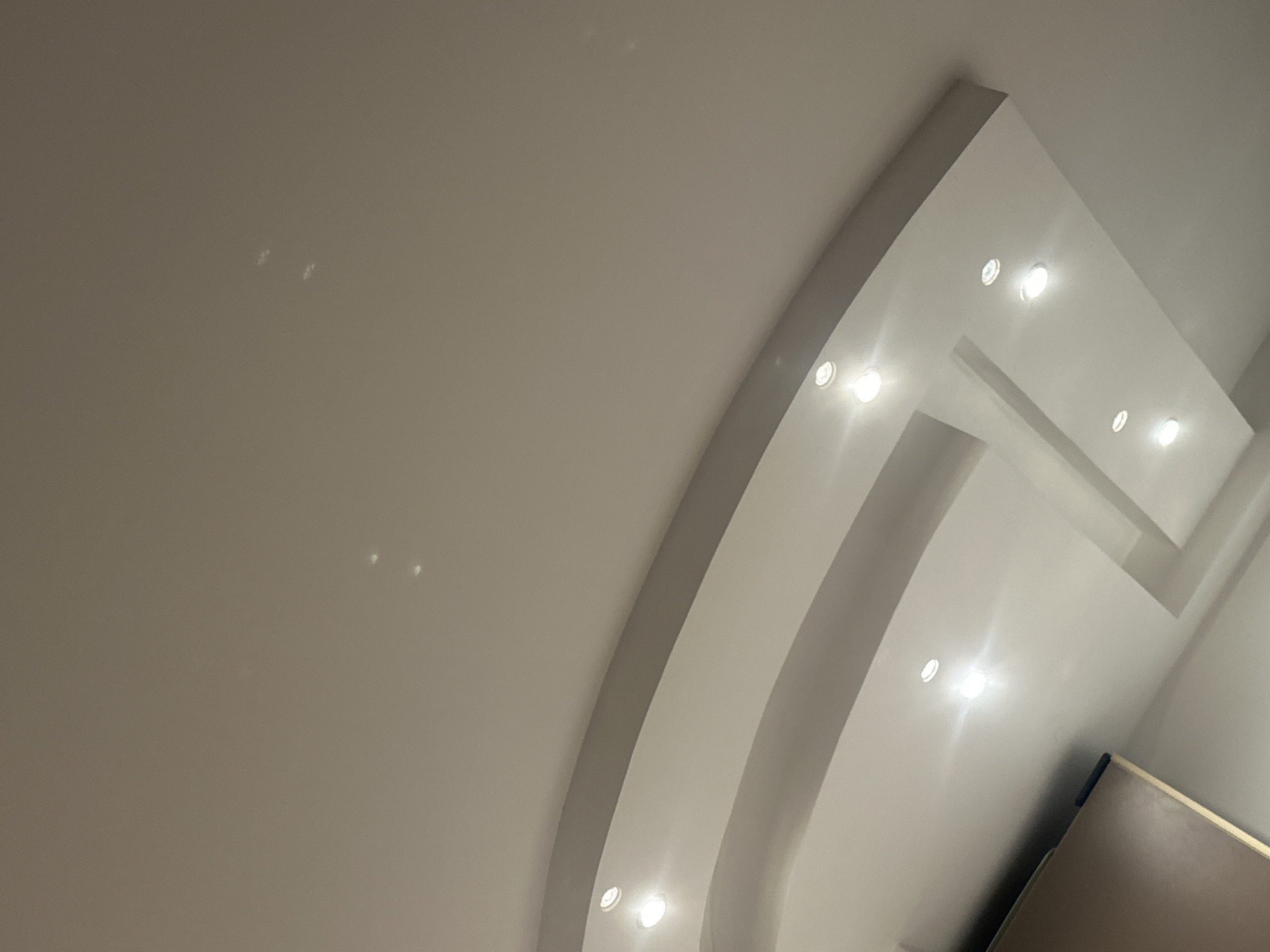 The image shows a section of a ceiling with recessed lighting. The ceiling has a complex, multi-tiered design with curved and rectilinear elements. The color of the ceiling is a light neutral tone, possibly white or off-white. The lighting fixtures are small, circular, and evenly spaced. A section of what appears to be a cabinet or furniture is visible in the lower right corner. The image does not reveal any information about the location being Sofia, Bulgaria or any people, subjects, actions, or interactions.