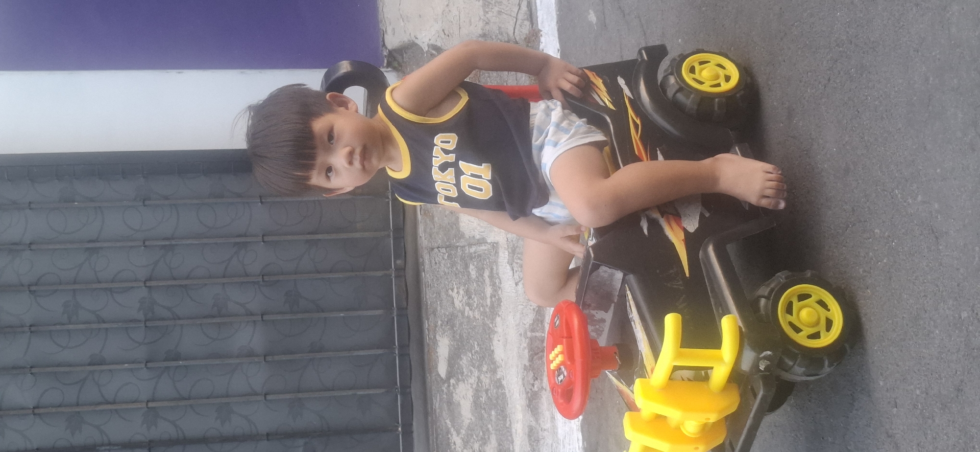 A young East Asian child is sitting on a black toy jeep with yellow accents, parked outdoors on an asphalt surface. The child is wearing a dark blue tank top with yellow trim and white lettering that reads "TOKYO 01", and striped shorts. Their feet are bare and resting on the jeep's footrest. A red steering wheel is visible in front of the child. Behind the jeep, a gray metal gate with a floral pattern is visible, and to the right, a portion of a purple wall can be seen. The location is identified as Desa Gagaksipat, Indonesia.