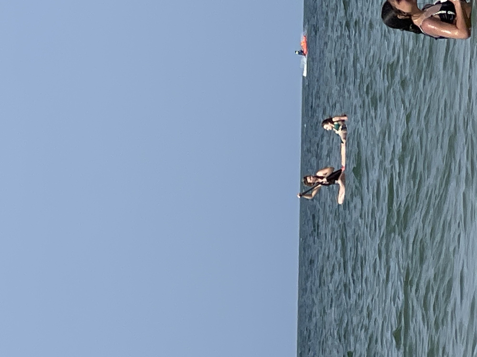 Two young women are paddleboarding on a calm, blue ocean under a clear sky. The woman in the front is sitting and holding a paddle. The woman behind her is lying down on the board. In the background, there is a small red and white object with a person on it, possibly another watercraft. In the foreground, a third young woman is visible, partially submerged in the water. The scene appears to be a recreational setting on a sunny day.
