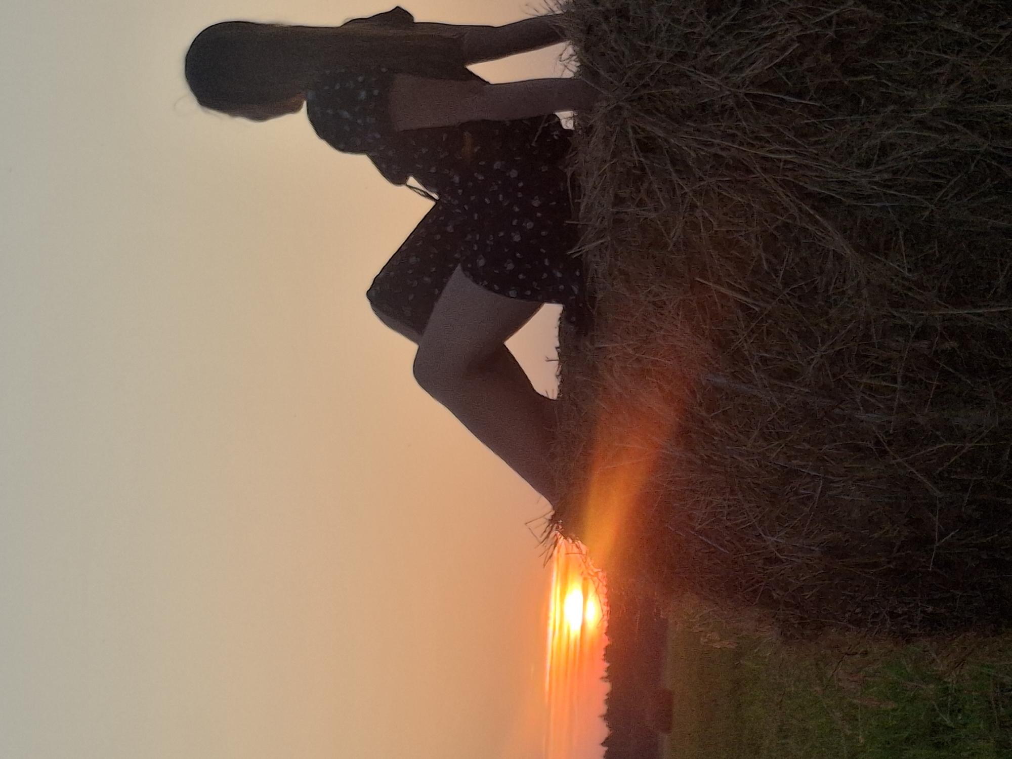 The image shows a silhouette of a young woman sitting on a haystack at sunset. She is wearing a dark dress with a floral pattern and is facing away from the camera, looking towards the horizon. The sun is setting, casting a warm orange glow across the sky and the surrounding field. In the background, a line of trees can be seen against the vibrant sunset. The haystack is large and appears to be in a rural setting, possibly on the outskirts of Perevoznoe, Russia. The overall mood of the image is peaceful and serene, with a hint of melancholy.
