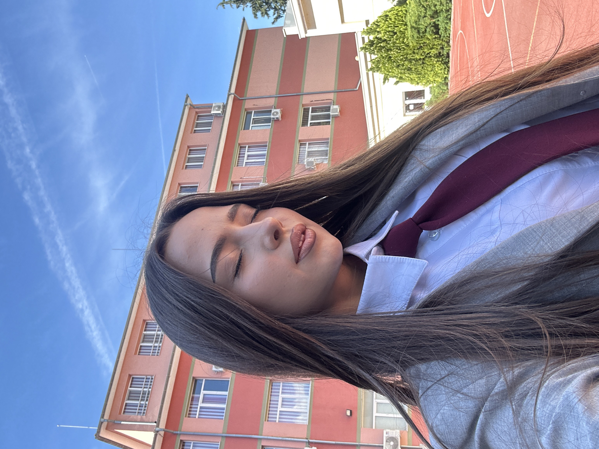 The image depicts a young woman with long, dark brown hair, closed eyes, and full lips. She is wearing a light grey blazer over a white collared shirt with a dark red tie. Behind her is a tall, multi-story building with alternating vertical stripes of red and salmon, and numerous windows. An air conditioning unit is visible on one of the building's exterior walls. The sky is blue with wispy contrails. A portion of a green tree and a red sports court can be seen in the upper right corner. The location and city are not discernable from the image.