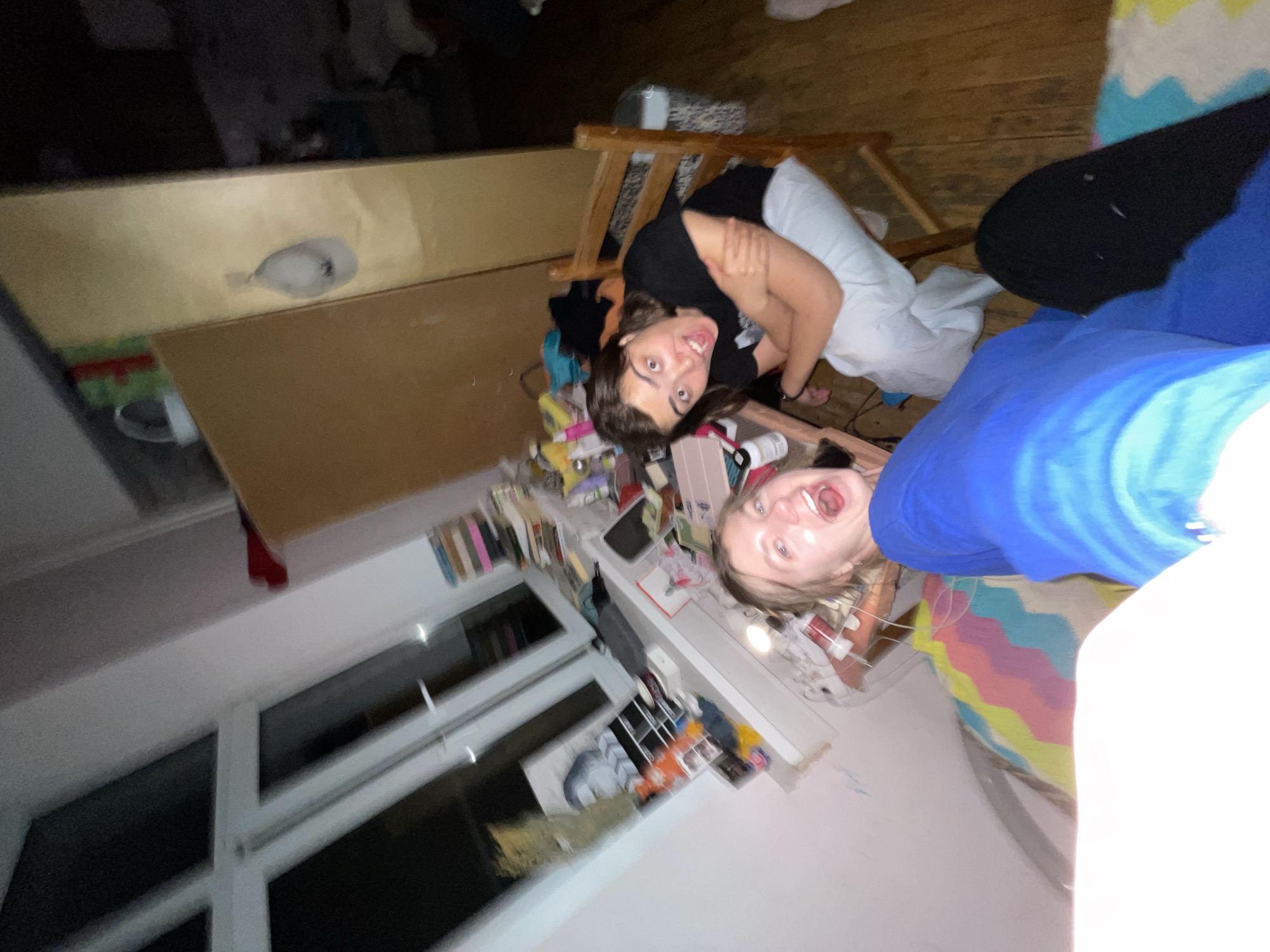Two young women are posing for a photograph in an indoor setting. The location appears to be a bedroom with a cluttered shelf containing books, art supplies, and other miscellaneous items. A window is visible in the background. One woman is wearing a black shirt, and the other is wearing a blue shirt. The women are smiling and looking directly at the camera. There is no evidence to suggest the location is Moscow, Russia.