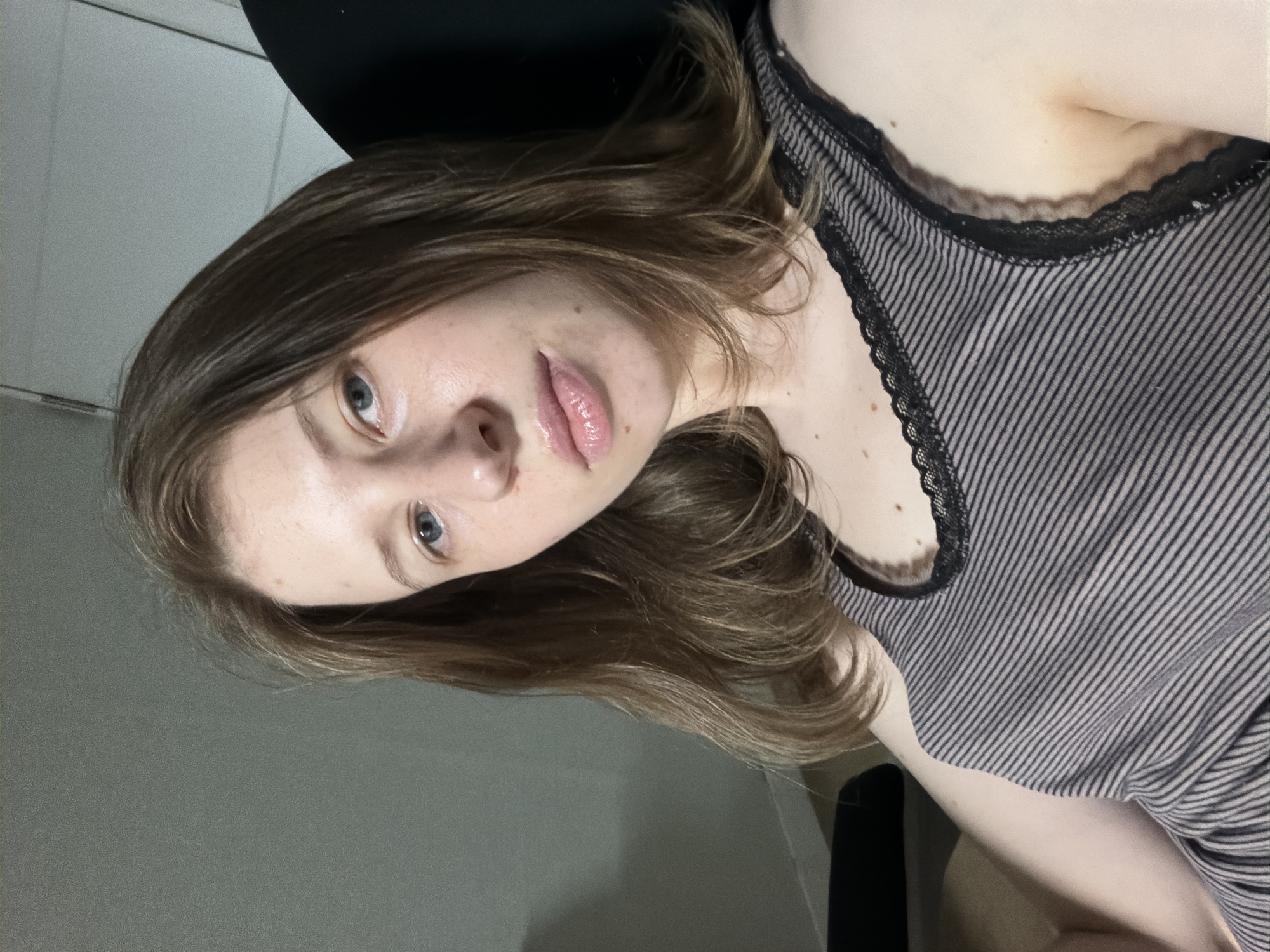 A young Caucasian female is seated, facing the camera. She has light brown hair that falls to her shoulders and blue eyes. Her lips are glossy. She is wearing a grey and black striped tank top with black lace trim. The background consists of a light grey wall and a portion of a black chair. The scene appears to be indoors.