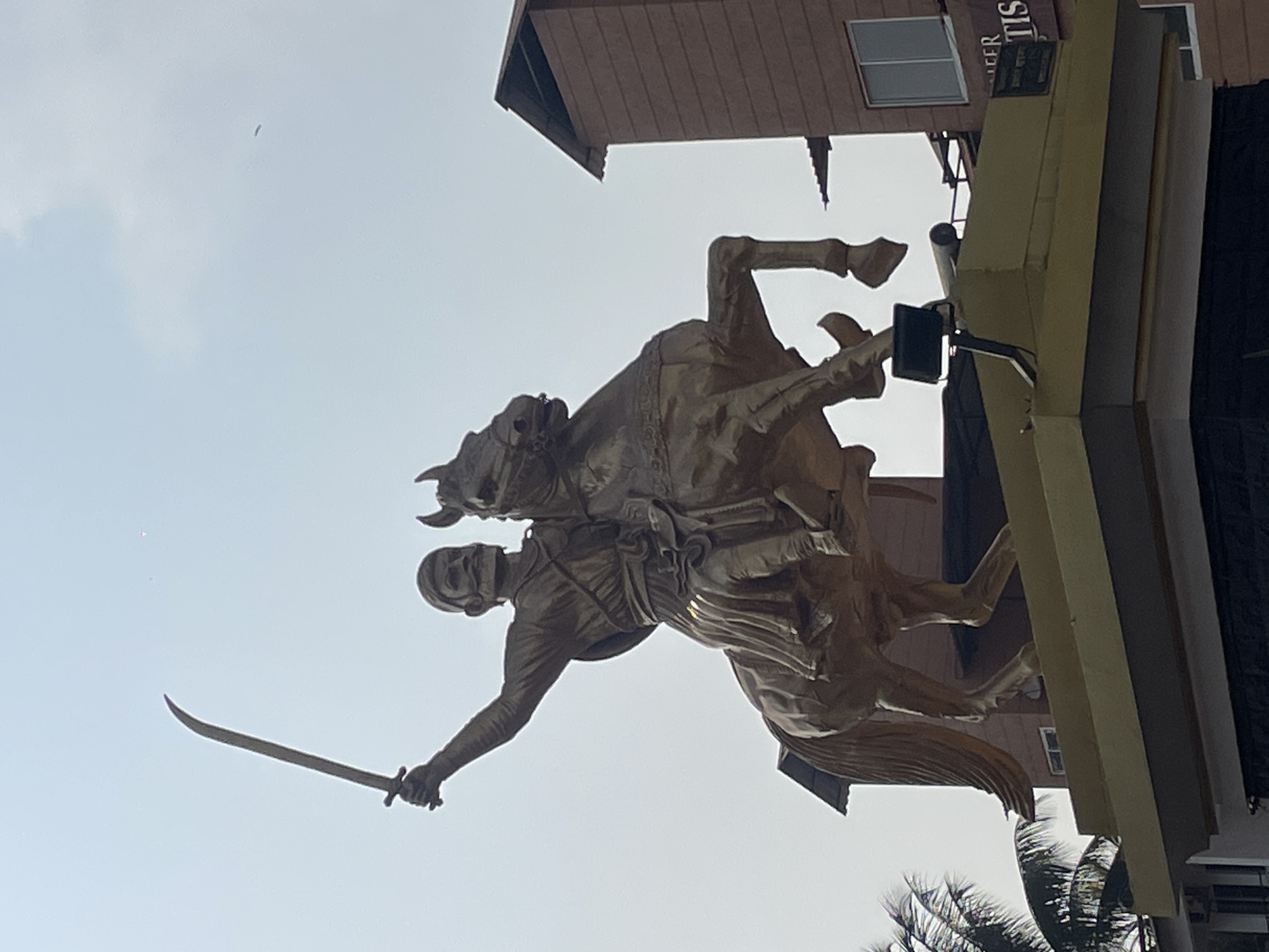 The image features a bronze statue of a man on horseback, holding a sword aloft. The statue is positioned in front of a building with a brown roof and yellow awnings. A sign with some words is barely visible behind the yellow awnings. The sky is visible in the background, with a small object that appears to be a bird flying high in the sky. The image is taken at an angle, making the statue and building appear tilted. The photograph was taken in Oxel, India.