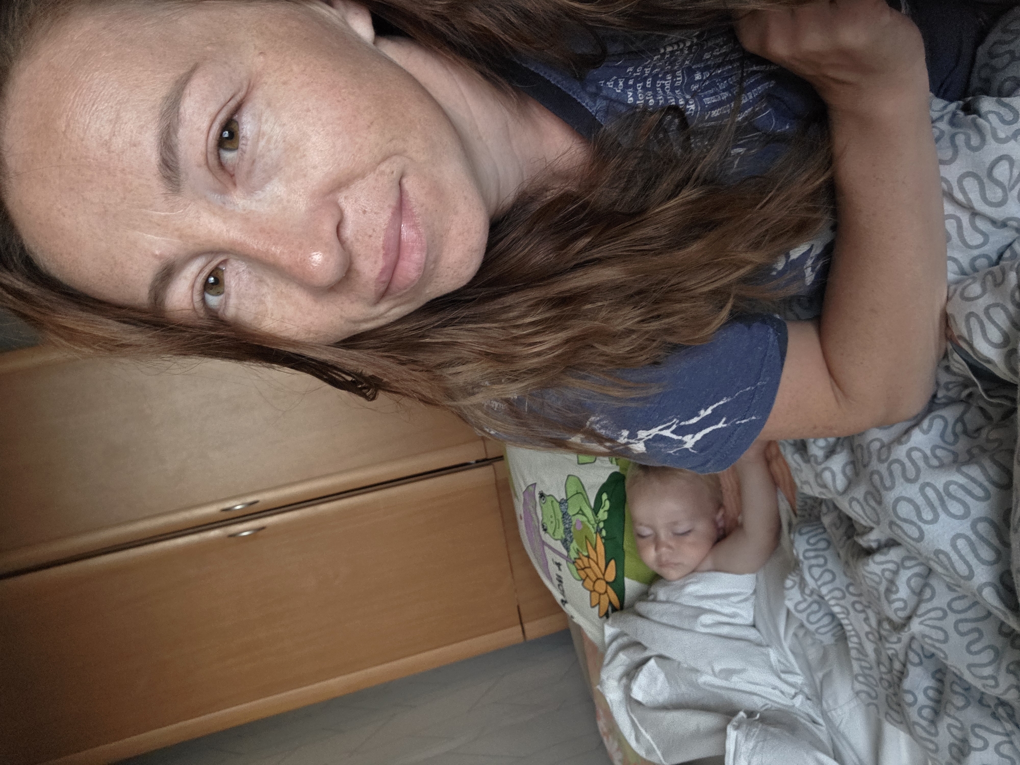 An adult female, appearing to be in her late 20s to early 30s, with auburn hair and a light complexion with freckles, is shown in a close-up selfie. She is wearing a blue t-shirt. She is in a room with a wooden wardrobe visible in the background. To her left, a baby is sleeping peacefully in a bed, covered with a white blanket and resting on a pillow with a frog illustration. The bedding has a gray wavy pattern. The overall scene suggests a domestic setting within a room in Буренка, Russia.