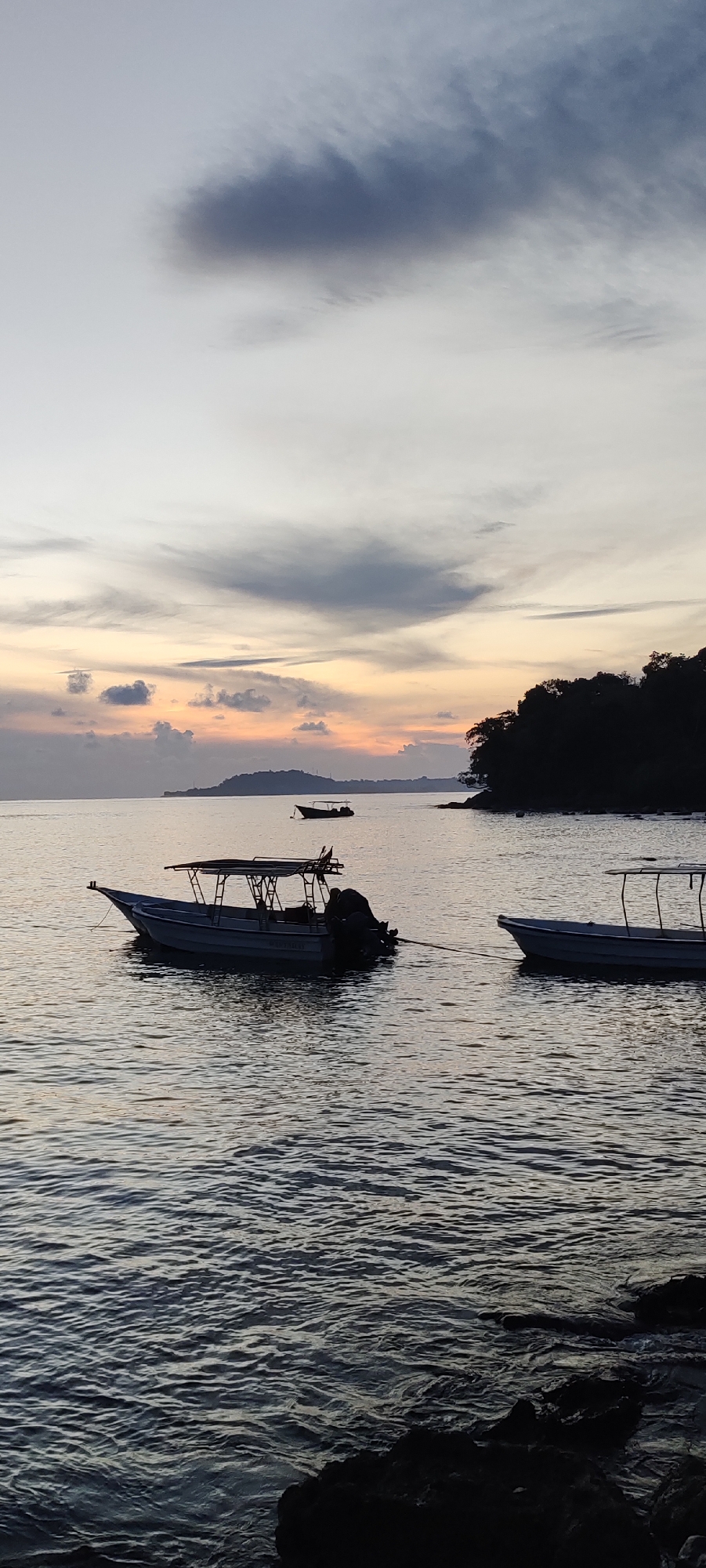 This image depicts a sunset scene at a coastal location in Sabang, Indonesia. The sky is a gradient of pale blue at the top, transitioning to hues of orange and yellow closer to the horizon, with scattered clouds casting shadows. In the foreground, the dark, rippling water of the ocean is visible, with rocks along the shoreline. Two motorboats are moored in the water. The boat on the left is larger, with a canopy and an outboard motor. The boat on the right is smaller and appears to be a fishing boat. A silhouetted tree line of a nearby island or coastline forms a dark mass on the right side of the image, and a distant, smaller island is visible on the horizon. The overall mood is serene and tranquil, characteristic of a tropical sunset.