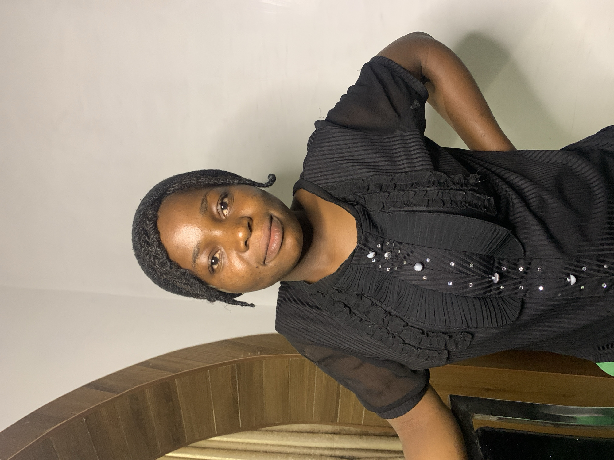 A dark-skinned young woman, wearing a black ruffled shirt adorned with sequins and beads, stands with her hands on her hips, looking directly at the camera. She has her hair styled in braids that fall to her sides. The background features a wooden archway with curtains visible through it, and a plain wall. There is no visible indication of the city of Jos, Nigeria.