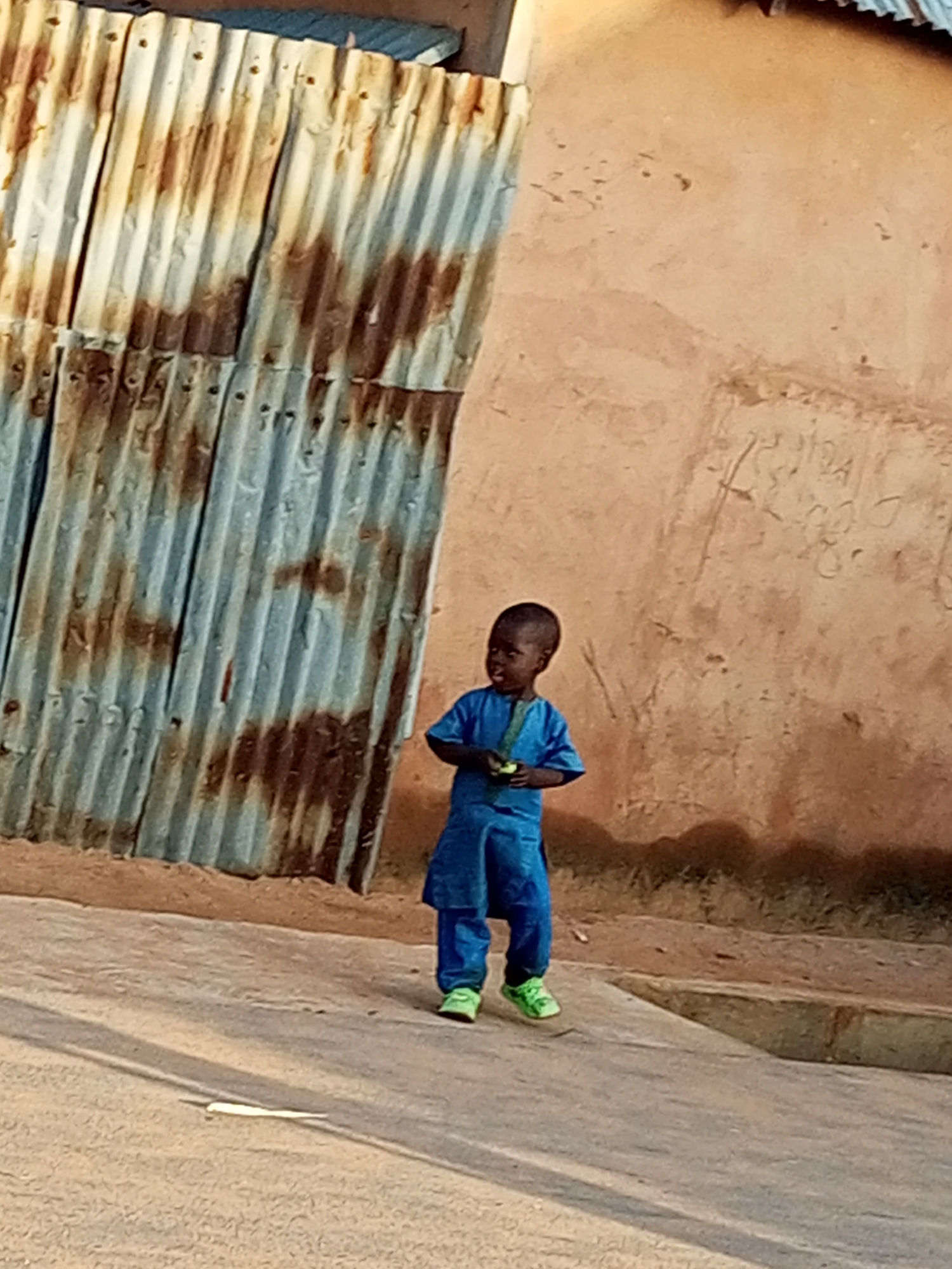 In Gombe, Nigeria, a young child stands on a dusty, paved surface. The child, dressed in a blue outfit, holds a light-colored object in their hands. To the left of the child is a corrugated metal wall, showing signs of rust and weathering. Behind the child is a textured, tan-colored wall, possibly made of mud or concrete, with faint markings or graffiti. The ground shows shadows cast from the surroundings.