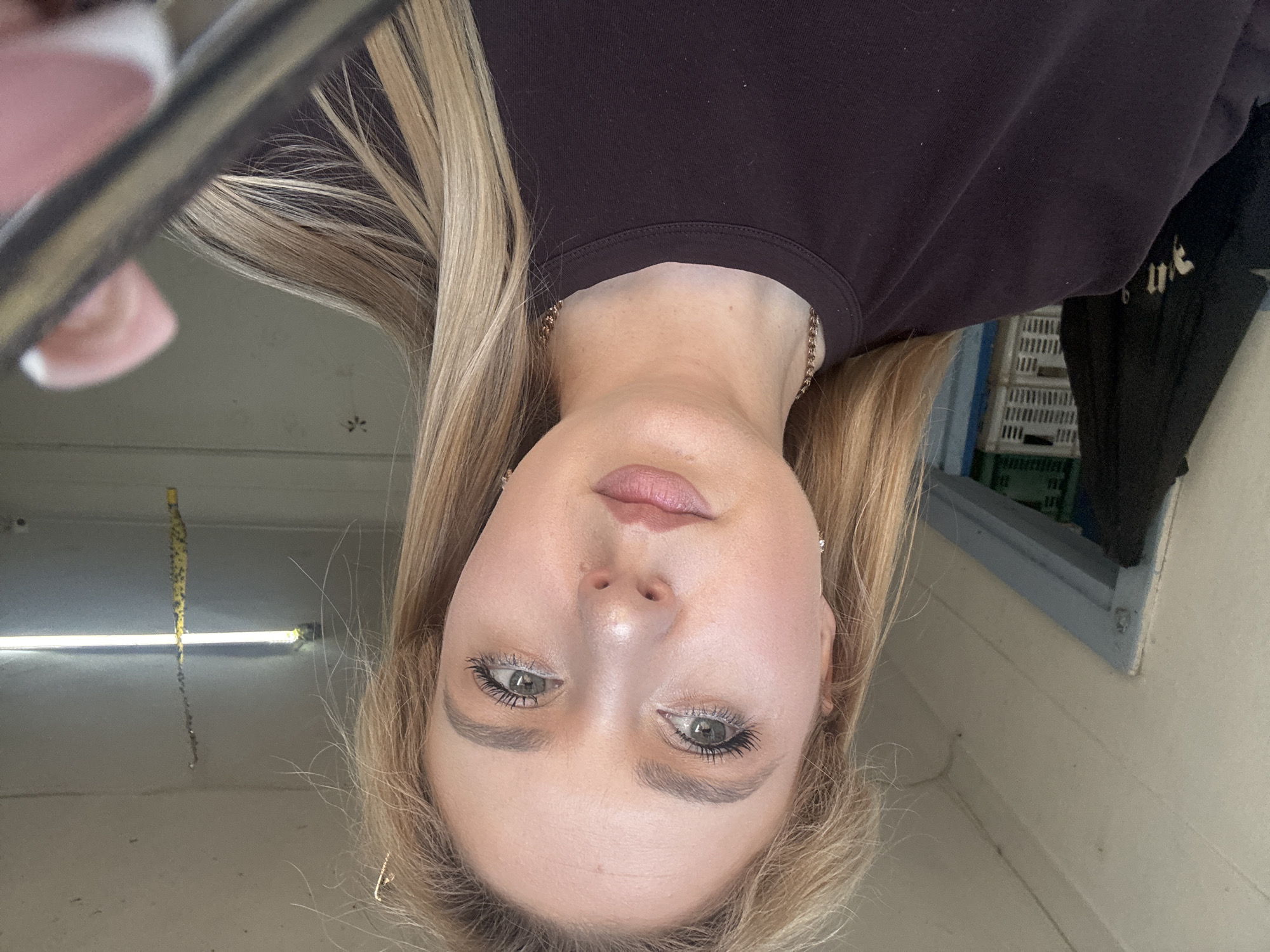 A young adult Caucasian woman with blonde hair is shown from the chest up, taking a selfie in what appears to be a storage or workshop area. She is wearing a dark purple crew neck t-shirt and a thin gold chain necklace. Her makeup includes mascara, eyeshadow, and lipstick. In the background, to her left, is a partially visible doorway with a blue frame and a black jacket hanging on a hook. Beyond the doorway are stacked white plastic crates. To her right, the ceiling has a fluorescent light fixture and a yellow and black striped object hanging down. The overall lighting suggests an indoor, possibly industrial or utilitarian space.