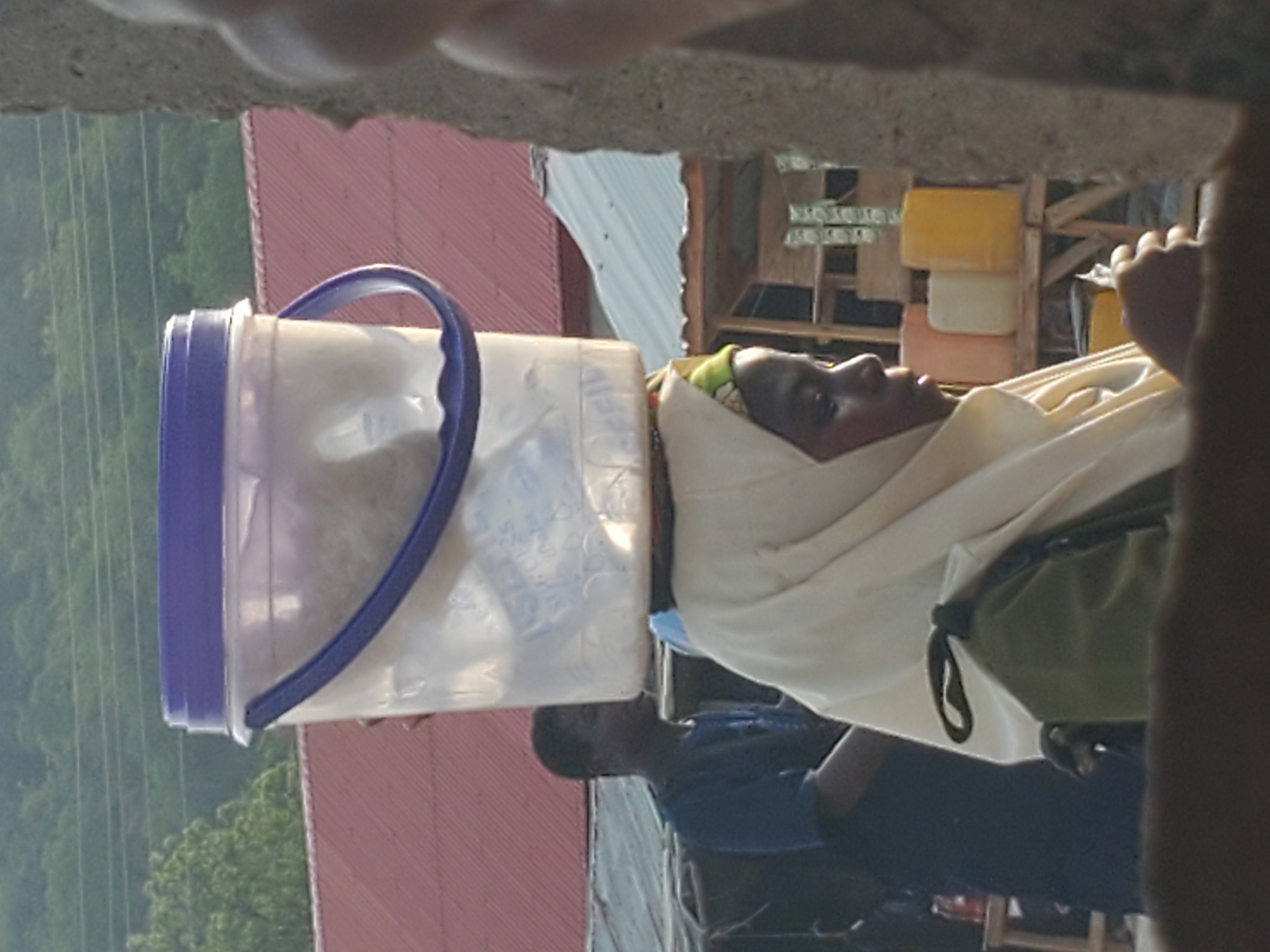 The image displays a woman expertly balancing a large, clear plastic bucket on her head, likely in an outdoor setting in Mararaba, Nigeria. The woman, with dark skin, is positioned on the right side of the frame, looking slightly upward. She wears a light-colored head covering, possibly a shawl or hijab, over a patterned headscarf. A dark green shoulder bag is also visible. The transparent bucket has a blue lid and handle and is filled with a light, granular substance, possibly grain or dry goods. Partially visible below the woman is another person, possibly a man, wearing a blue shirt.
In the mid-ground, a building with a red corrugated metal roof is prominent. Further in the background to the left, lush green trees and what appear to be power lines suggest an outdoor environment. To the right in the background, a wooden structure displays several colorful plastic containers – yellow, cream, and pink – possibly goods for sale at a market stall. The photograph is taken from a slightly elevated and partially obscured vantage point, with a blurry concrete or stone element framing the top. The lighting indicates daytime, with soft illumination and no harsh shadows. Faint, unreadable text is visible on the plastic bucket and possibly on distant signs. The woman's expression is neutral, reflecting concentration on carrying her load.