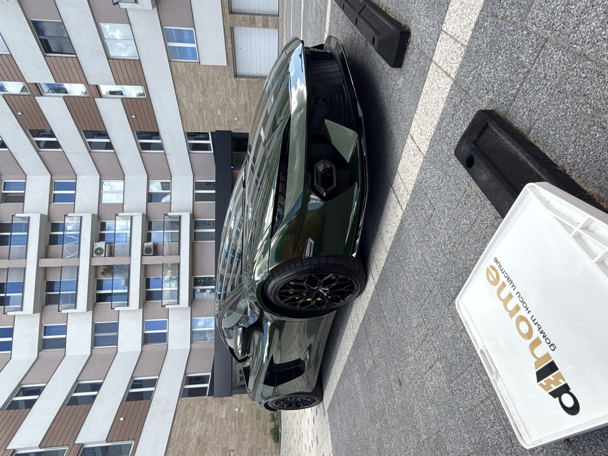 The image shows a dark green Lamborghini parked on a paved surface next to a modern building with a facade of beige and brown horizontal stripes, featuring many windows that reflect the sky. The location appears to be an urban area, specifically identified as Sofia, Bulgaria, based on the Cyrillic lettering "домът носи щастие" on a nearby white plastic bin, which translates to "home brings happiness". Two black rectangular parking bumpers are visible near the car. The scene seems to be taken on a bright, sunny day.