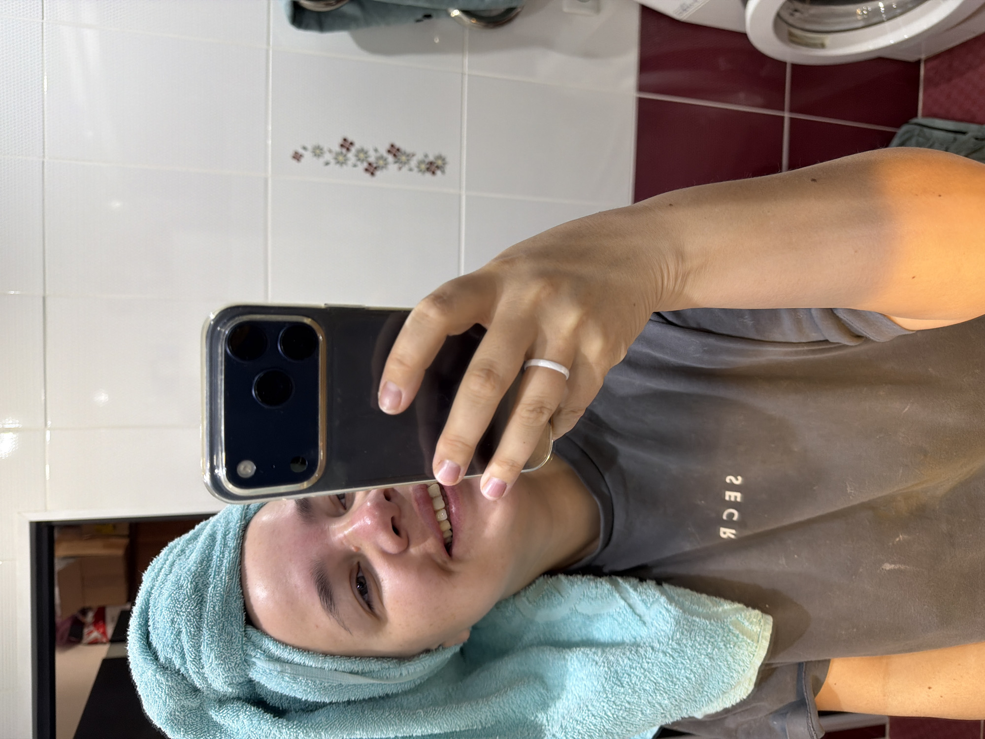The image captures a close-up mirror selfie of a person, likely a woman, in a bathroom setting in Chaikovsky, Russia. The subject is seen from the chest up, smiling gently at the camera. A light blue towel is wrapped around her head, indicating she may have just washed her hair or is about to.
She holds a dark-colored smartphone in her right hand to take the picture; the phone's rear camera array and a clear protective case are visible. On her right ring finger, she wears a simple white ring. Her expression appears content and relaxed. She is wearing a grey t-shirt, on which white, mirrored text is visible. The text, appearing as "ЯЭCК" in the image, would read as "КCEЯ" or "RSEK" when un-mirrored.
The background consists of white square wall tiles, some featuring small decorative floral patterns in shades of brown and white. Below these are reddish-brown square tiles. A part of a white appliance, resembling a washing machine with a circular door, is visible in the upper right section of the frame. The lighting seems bright, possibly artificial, highlighting her skin with a warm tone on her exposed arm.