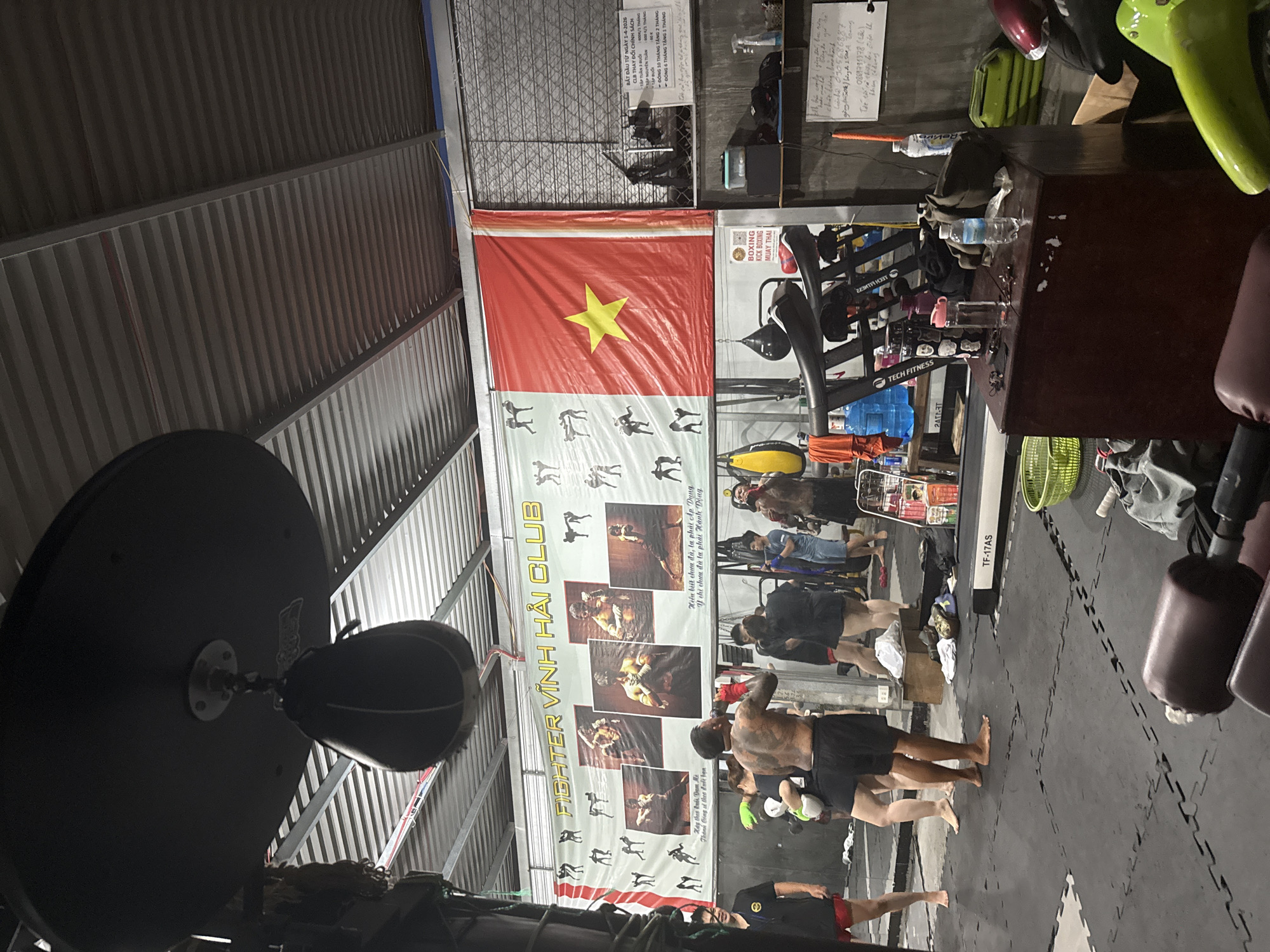 This image depicts the interior of a gym in Bac Nha Trang Ward, Vietnam, likely a martial arts training facility. The scene is captured from a slightly low angle, looking towards the front of the gym.
Dominating the background is a large banner with "FIGHTER VINH HAI CLUB" written in yellow capital letters. The banner also features images of martial artists in various poses and a large Vietnamese flag with a yellow star on a red background. To the right of the banner, a metal grate covers a window or wall section, and a sign is attached, listing class schedules and prices.
In the foreground and mid-ground, several individuals are present. One man, shirtless and with extensive tattoos, is standing and drinking from a water bottle. He is wearing red boxing shorts and wrist wraps. Other people are visible in various states of activity, some appearing to be training or resting. There are several pieces of gym equipment, including a speed bag hanging from the ceiling, treadmills, and what appear to be weights or training dummies.
The gym floor is covered with black interlocking mats. The ceiling is constructed of corrugated metal and exposed beams, a common feature in this type of facility. Various items are scattered around, including water bottles, gym bags, and personal belongings. The overall atmosphere suggests a functional, no-frills training environment.