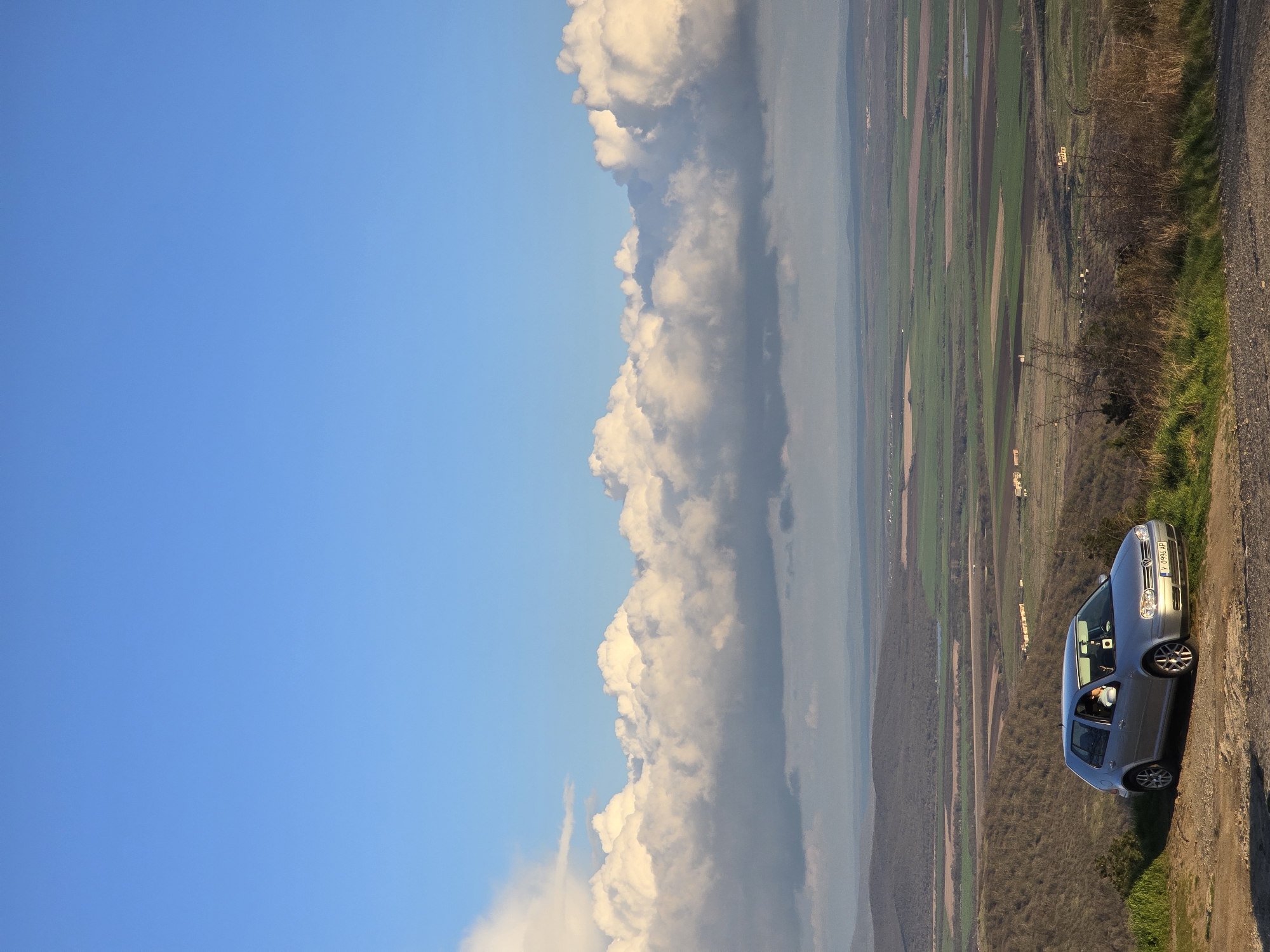 A silver sedan is parked on a grassy verge beside a dirt road in Pobeda, Bulgaria, offering a panoramic view of the landscape. The sky, largely clear and blue, transitions into a dense bank of cumulus clouds along the horizon. Below, alternating strips of cultivated fields and forested areas stretch into the distance. A person is visible inside the car.