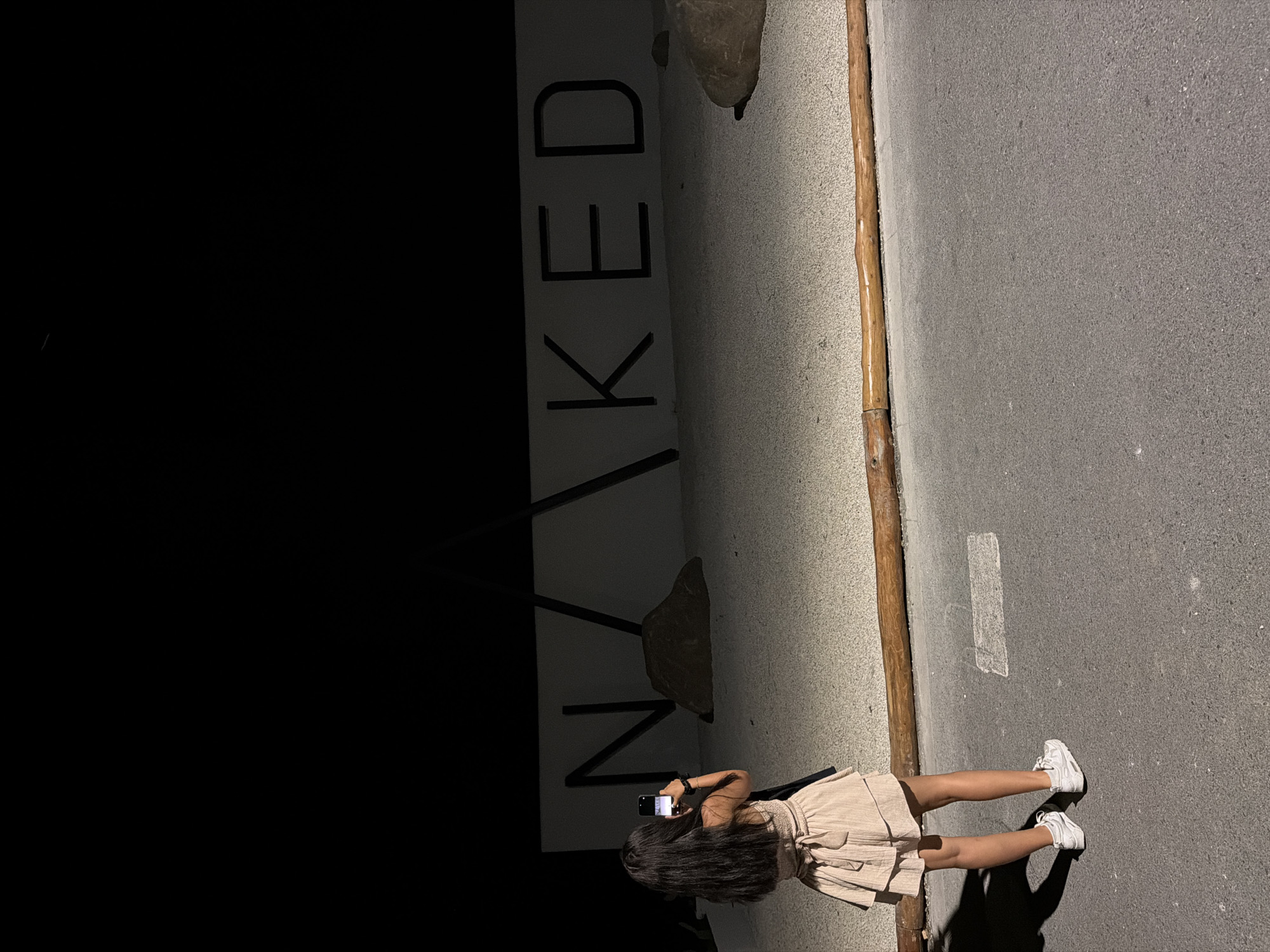 A woman stands on an asphalt road next to a wall with large letters spelling "NAKED". She is wearing a light-colored skirt and white sneakers and is holding a phone up to take a picture of the wall. The lighting suggests it is nighttime or very early morning. The wall appears to be made of a rough, textured material, and the letters are dark and bold. There's a rough wooden post running vertically along the edge of the wall. The overall scene feels like an urban or semi-urban environment, possibly near a business or art installation. The woman's posture suggests she is actively engaged in capturing the moment.
