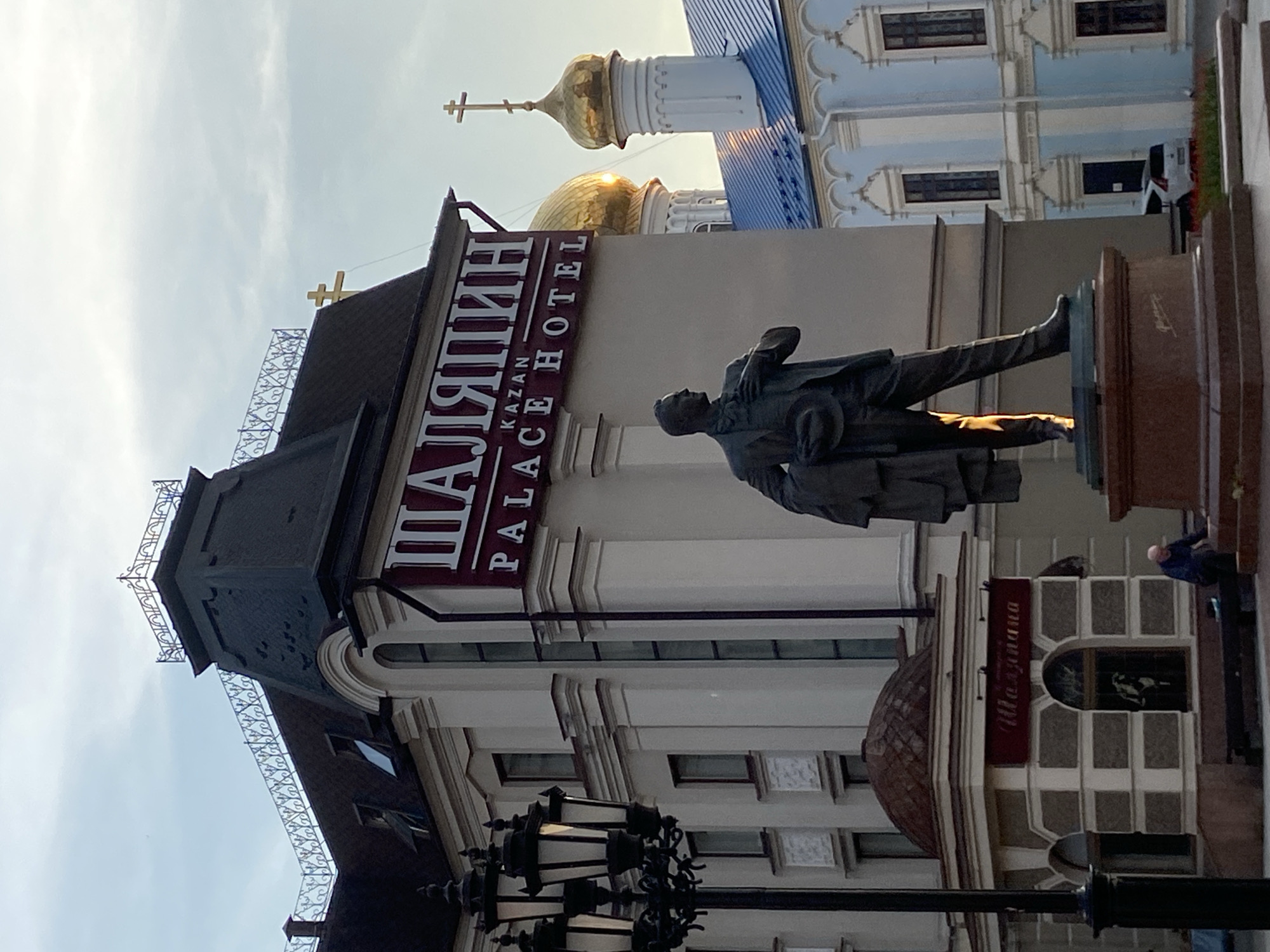 The image depicts a bronze statue of a man in a suit and hat. He is standing on a pedestal in front of a building with a sign that reads "Shalyapin Kazan Palace Hotel" in English and Russian. Behind the hotel, the domes of a blue and white church can be seen. The image appears to be taken in Kazan, Russia. Other elements of the scene include ornate streetlights, another building labeled "Ilzazziana," and partial figures of people in the background.