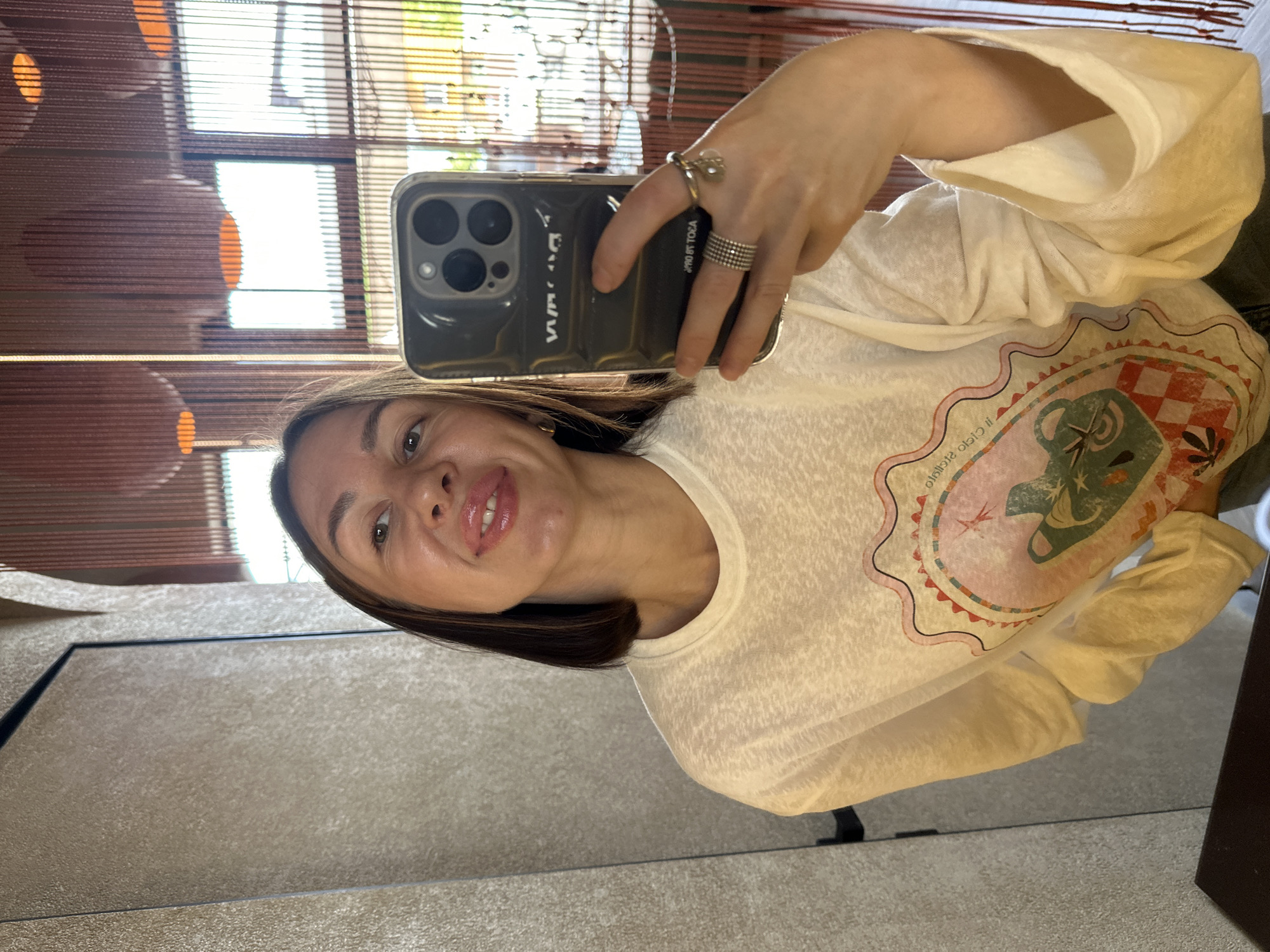An adult female is depicted taking a mirror selfie in an interior setting located in Усады, Russia. She holds a dark-colored smartphone, consistent with an iPhone Pro Max model, encased in a clear protective cover, in her right hand. Reflected text "VWA" and "ЯJOЯ OT TОEА" is visible on the phone case. Her right hand displays multiple rings: a wide, multi-band ring on the ring finger and a ring with a charm (appearing as a clear bead and metal element) on the middle finger. She has medium-length dark brown hair, light complexion, and is smiling slightly with glossy pink lips, revealing upper teeth. A small gold-colored hoop earring is visible on her left ear. She is wearing a light-colored, long-sleeved t-shirt with a central graphic design featuring a scalloped border, a green anthropomorphic creature, checkerboard patterns, and floral elements in shades of pink, green, orange, and white. The background, reflected in the mirror, shows a textured light-colored wall on the left. To the right, a decorative screen of numerous reddish-brown vertical strings or beads is present. Behind this screen, multiple round, orange-glowing light fixtures are visible, along with large windows revealing a brightly lit outdoor environment with green foliage. A dark, flat surface occupies the immediate foreground.