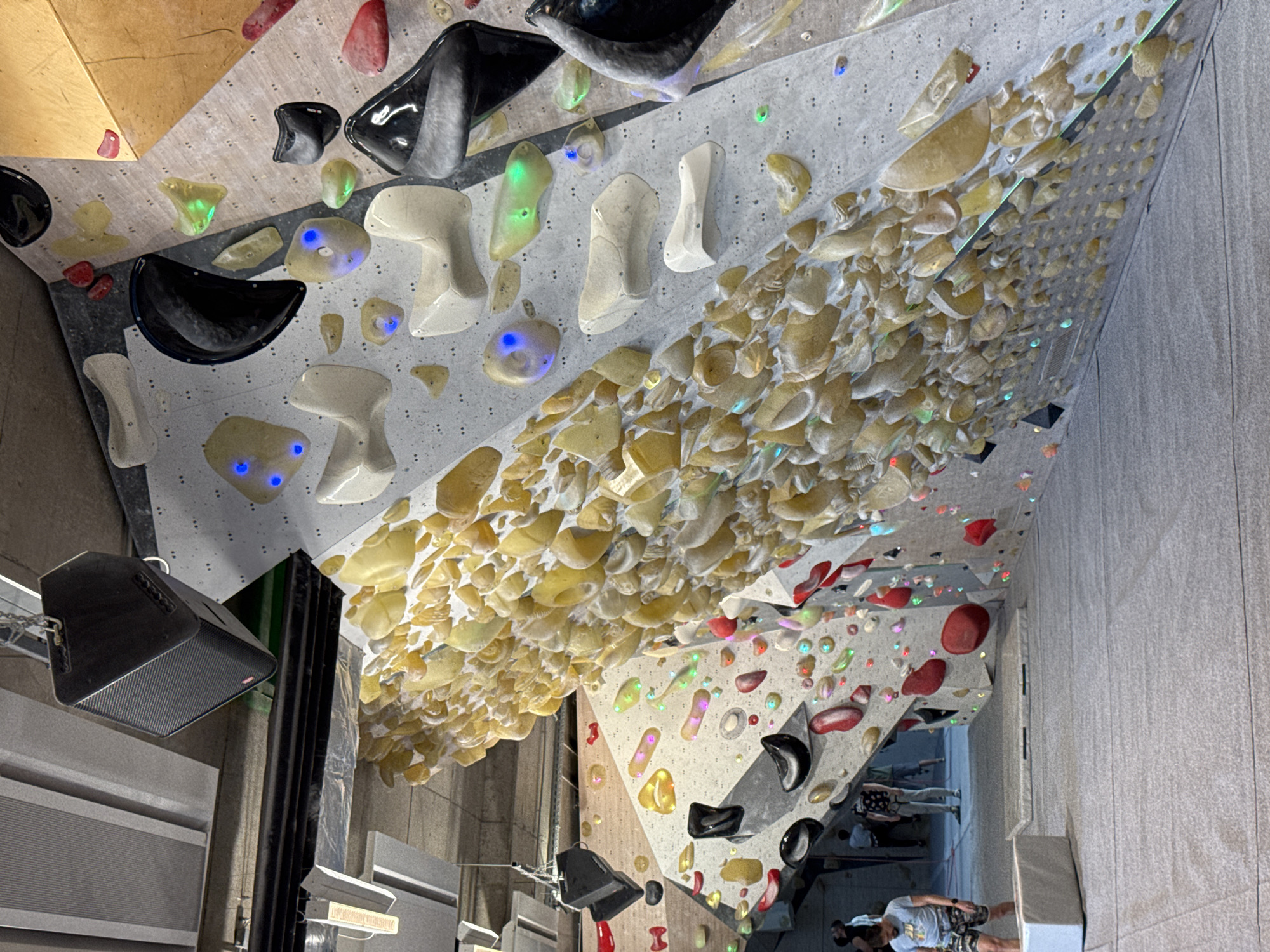 This image captures an interior view of an indoor bouldering gym, featuring a complex arrangement of climbing walls and holds. The perspective looks up and across the climbing structures, which are covered with a dense variety of grips.
The walls are predominantly light gray, with some sections showing a wooden or concrete texture. A large overhang area is covered extensively with numerous small, yellowish-beige textured holds. On other wall sections, larger and more distinct climbing holds are visible in various colors, including black, white, red, and green. A notable detail is the presence of translucent holds that are illuminated with internal LEDs, glowing either blue or green, suggesting an interactive climbing system for specific routes.
In the lower left of the frame, a black, angled speaker is mounted to a beam, with metallic grating or panels visible below it. Towards the bottom right and in the background, several individuals are present, indicating the gym is in use. One person, wearing a light-colored t-shirt and patterned shorts, stands on what appears to be a crash mat, looking towards the walls. Other people are less clearly defined in the background.
The lighting is artificial, emphasizing the various holds, particularly those that are LED-illuminated. The setting is clearly an active climbing environment, likely during a bouldering session. No discernible text is visible on any objects or surfaces. Given the context, the location is a bouldering gym in Sofia, Bulgaria.