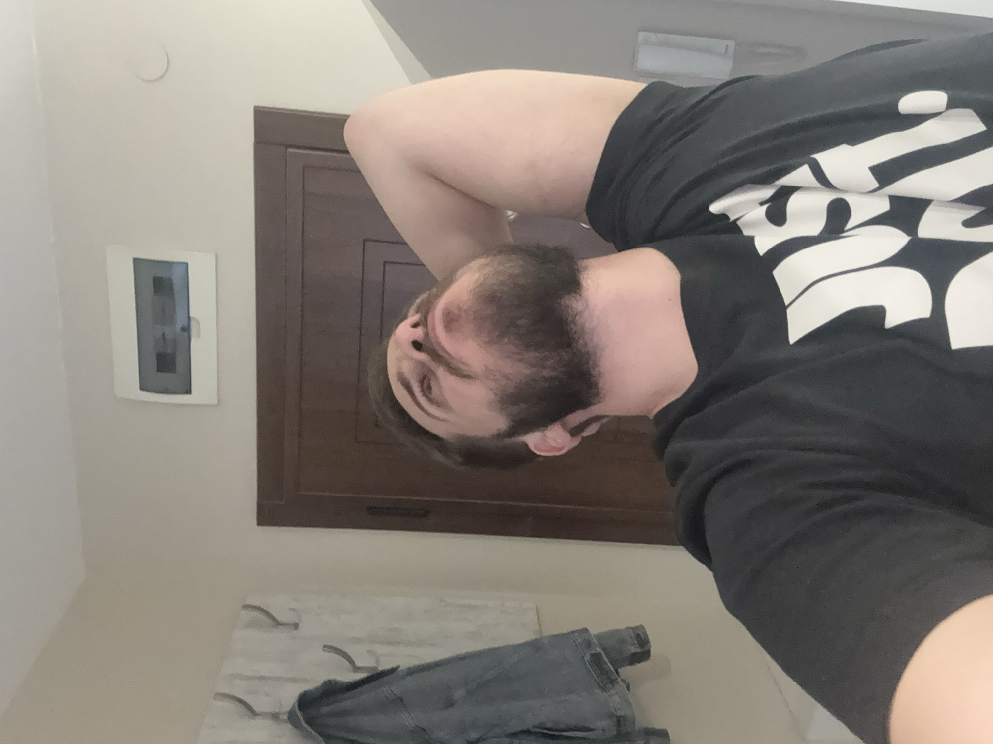 A selfie-style image features a person with a beard wearing a black t-shirt with white writing. In the background, there's a wall with a small, rectangular electrical box. A wooden panel with a rectangular frame is situated to the right of the electrical box. Below the person, a piece of clothing appears to be draped over hooks on a surface with a light marble effect. The location in the city of Sofia, Bulgaria is not discernible from the available visual information.