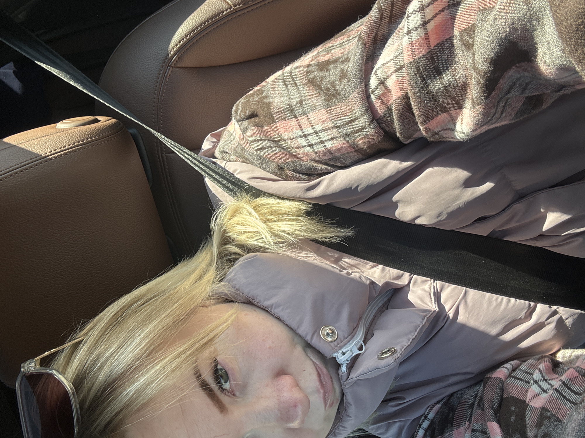 A blonde woman is in a car wearing a pink puffer jacket and a plaid shirt, restrained by a seatbelt. She has a pair of sunglasses. The car's interior features brown leather seats. The location and city cannot be determined from the image.