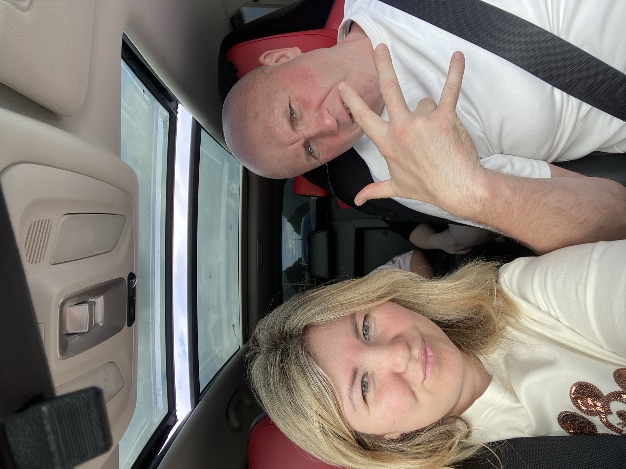 This is a selfie taken from inside a car, with the subjects positioned in the front seats. The image is oriented vertically. On the left, a woman with blonde hair is smiling at the camera. She is wearing a light-colored t-shirt decorated with sequined Mickey Mouse heads. On the right, a bald man with blue eyes is making a "rock on" hand gesture. He is wearing a white t-shirt and appears to be smiling or smirking.
The interior of the car features red and black seating, a black seatbelt, and a panoramic sunroof that reveals an overcast sky. The lighting suggests it is daytime. The scene appears to be set in a rural or suburban area, possibly in or near Новая Казмаска, Russia, given the context. The overall mood is casual and friendly. There is no visible text in the image.