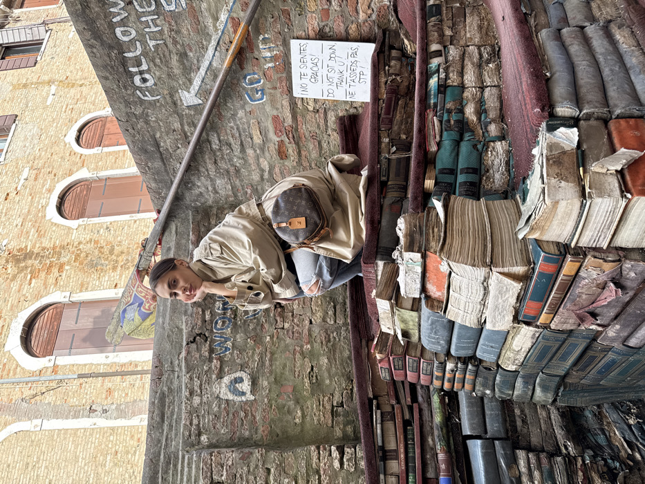 An outdoor, eye-level shot captures a woman posing on a unique staircase constructed from stacks of aged books at Libreria Acqua Alta in Venice, Italy. The woman, with dark hair pulled back, is in a crouching position on the middle tier of the book-steps, looking directly at the viewer with her left hand resting under her chin. She wears a light beige trench coat, light blue ripped jeans, and a brown Louis Vuitton monogram round crossbody bag. The steps themselves are made of numerous old, worn, and variously colored books, some appearing water-damaged, arranged on platforms covered with dark red material. Behind the woman and the steps, a weathered brick wall features graffiti including "FOLLOW THE S" and "GO UP" with an arrow. A metal handrail runs diagonally up the wall. To the right, a white sign is affixed to the wall, displaying "¡NO TE SIENTES, GRACIAS!", "DO NOT SIT DOWN, THANK U!", and "NE T'ASSSIEDS PAS, STP!" in black text. In the upper background, arched windows with dark shutters are visible on a brick building.