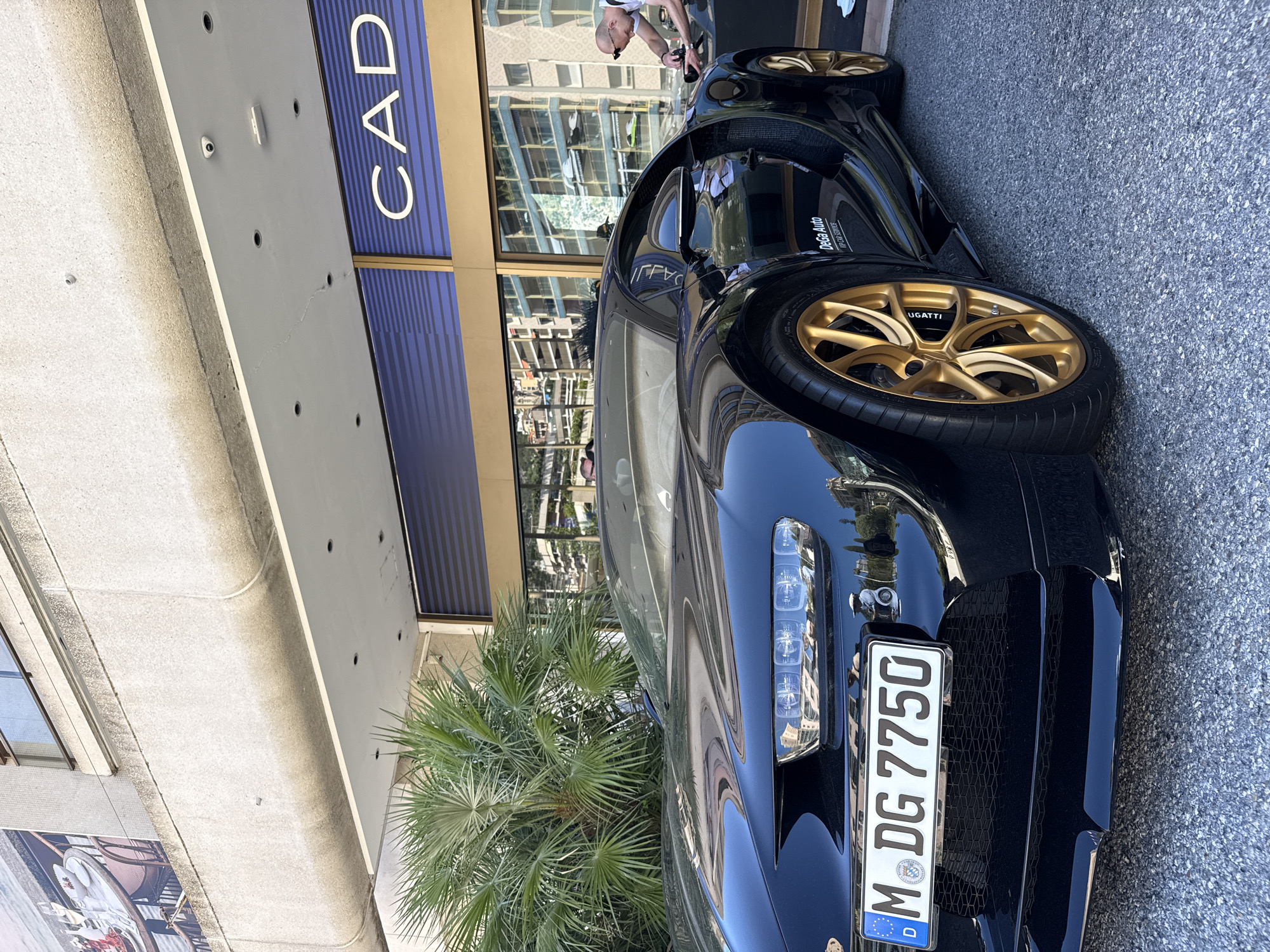A black Bugatti Chiron is parked in front of a building with a sign that reads "CAD" in large white letters. The car has gold rims and a license plate that reads "M DG 7750". A palm tree is visible behind the car. A man is taking a photo of the car. The scene appears to be in Monaco.