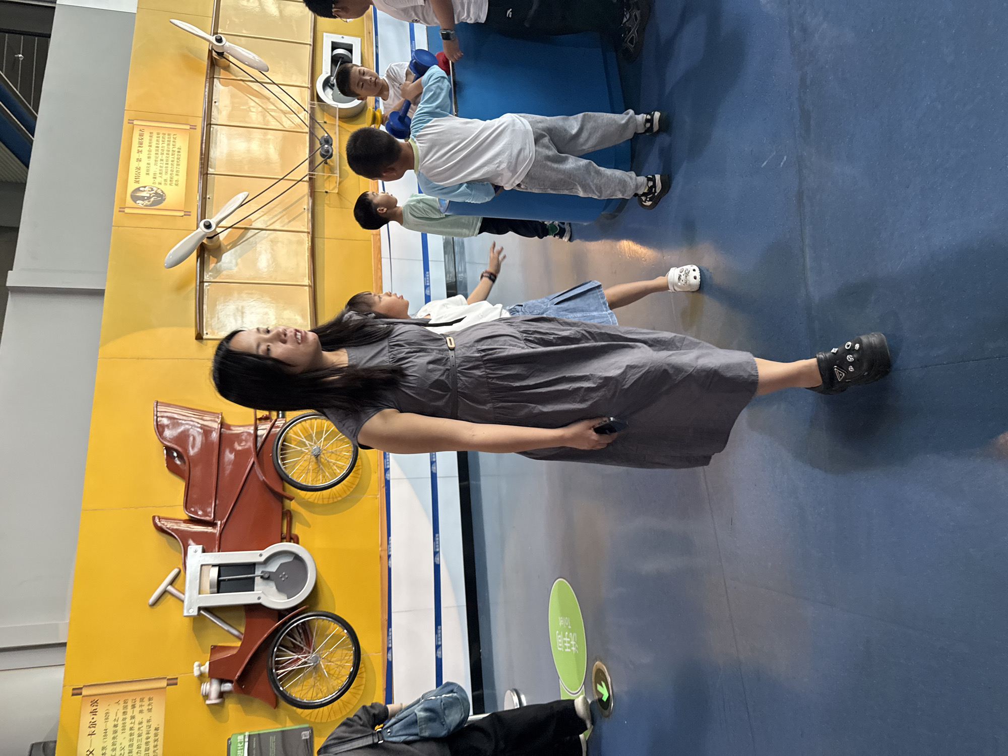 The photo shows an indoor scene in 江北城街道, China. A woman is standing on a blue floor, wearing a gray dress and black shoes. Several children are nearby, and behind her is a yellow wall with an exhibit featuring a vintage car and aircraft propellers. There are signs with Chinese characters on the wall as well. Another person is partially visible in the foreground, wearing black pants and carrying a blue bag.