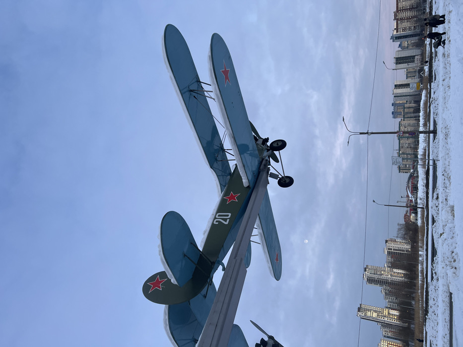 The image displays a biplane monument set against a sky and an urban landscape, presented in a portrait orientation as if rotated 90 degrees clockwise from its natural horizontal perspective.
In the prominent left and central portions of the image, a biplane is mounted on a display stand, angled upwards. The plane's wings are a light blue or teal color on their upper surfaces, and there's a noticeable layer of white snow accumulated on the top edges of both the upper and lower wings. The fuselage, partially visible, appears to be an olive green color. On the underside of the lower wing and on the tail fin, a red star with a white outline, characteristic of Soviet-era aircraft, is clearly visible. The number "20" is also distinctly marked in white on the side of the fuselage. The biplane's landing gear, featuring two wheels, is visible near the top right section of the plane.
The left side of the image is dominated by the sky, which is a clear, light blue with some scattered, wispy clouds. A faint, circular white object, likely the moon, is visible against the blue sky, indicating it's daytime.
The right side of the image reveals an urban environment covered in snow. Numerous tall, multi-story buildings, characteristic of modern high-rises or apartment complexes, line the distant horizon. The ground in the foreground and midground on the right is heavily covered in white snow, suggesting a winter season. There are two streetlights with curved arms visible. Near the buildings, small dark figures that appear to be people can be seen walking on the snow-covered ground, adding a sense of scale and life to the scene.
