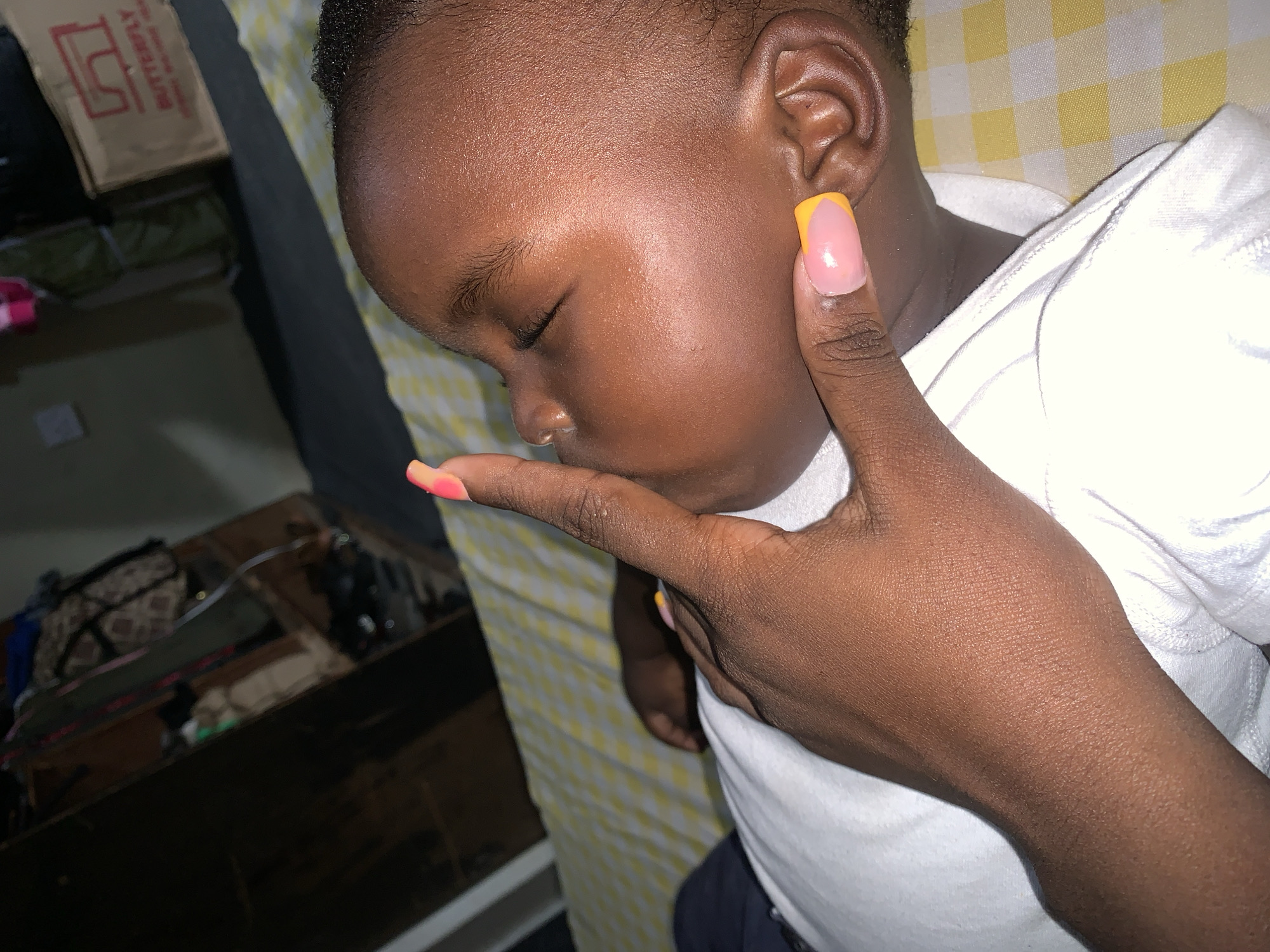 A close-up, high-angle shot shows the right side of a young child's face and head, with a person's hand gently touching the child's lips and ear. The child is lying down, eyes closed, with fair skin and dark hair. The person's hand has manicured fingernails painted with a pink and orange gradient. The child is wearing a white garment. The background shows a yellow and white checkered pattern, possibly a bedsheet, and some blurred items in the distance, including what appears to be a box with "BUTTERFLY" written on it. The overall scene is intimate and peaceful.
