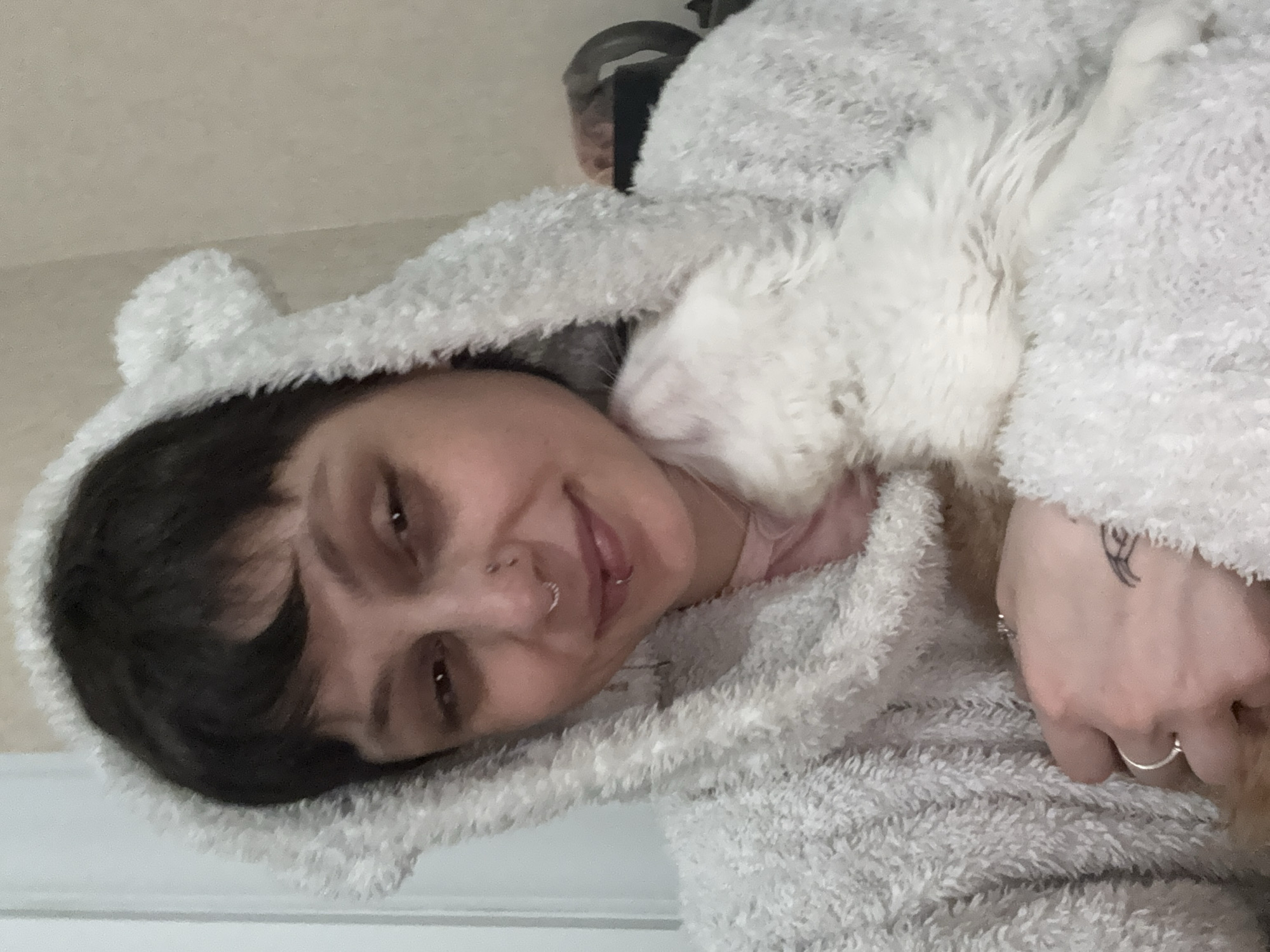 An adult female is captured indoors in Saint Petersburg, Russia. She wears a light grey, fluffy hooded garment featuring two rounded, ear-like protrusions, over a pink undergarment. Her short, dark hair includes bangs. Facial piercings consist of a silver septum ring, a silver nostril stud, and a silver labret ring on the lower lip. She exhibits a subtle smile and direct eye contact with the viewer. She is holding two felines: a white, fluffy cat is nestled against her left shoulder and neck, its head turned upwards and away from the camera, while a second, partially visible animal with orange-brown fur, consistent with a cat, is held by her left arm. Her left hand, adorned with two silver rings and a small dark tattoo on its dorsal surface, supports the animals. The background is a light-colored, out-of-focus wall, with a lighter vertical element on the left side, indicating an interior setting.