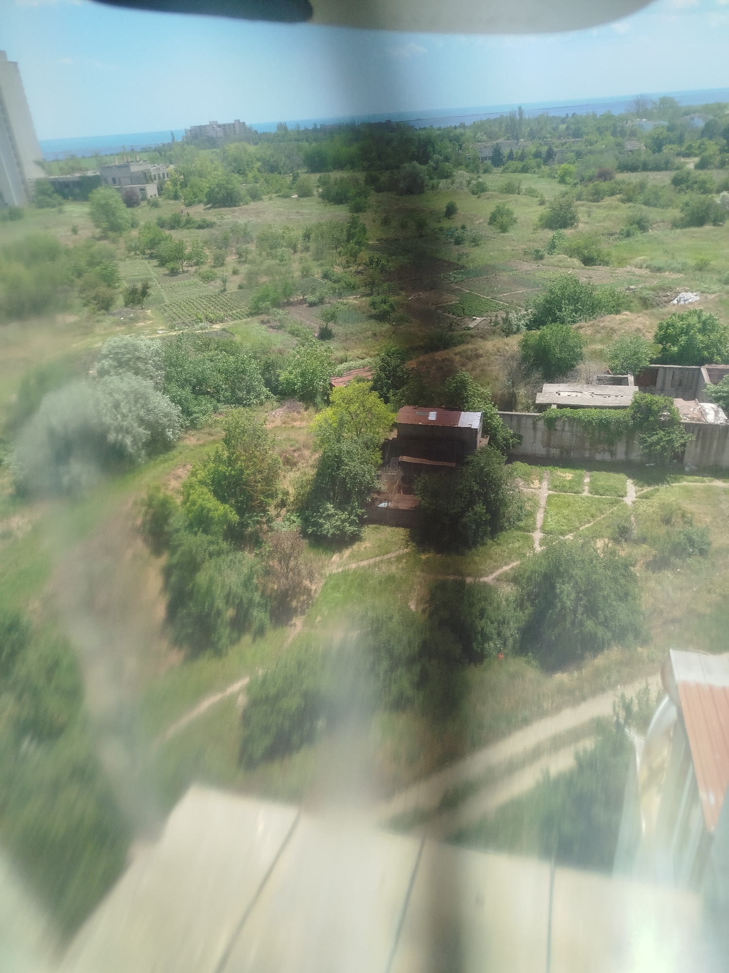This is an aerial view of a semi-developed landscape with a body of water in the distance. In the foreground, a light-colored surface, possibly a roof or balcony railing, takes up the bottom portion of the image. The middle ground features a grassy area with scattered trees and vegetation. A small, dilapidated structure with a rusty metal roof is visible in this area. Further back, several other derelict concrete buildings and structures are scattered amongst overgrown vegetation. Rows of cultivated plants or gardens are also visible in the mid-distance. In the far distance, a body of water, likely the sea or a large lake, meets the horizon under a clear blue sky. A tall, light-colored building is visible on the left side of the frame in the background, suggesting a coastal urban or semi-urban environment. The overall impression is one of abandonment or underdevelopment.