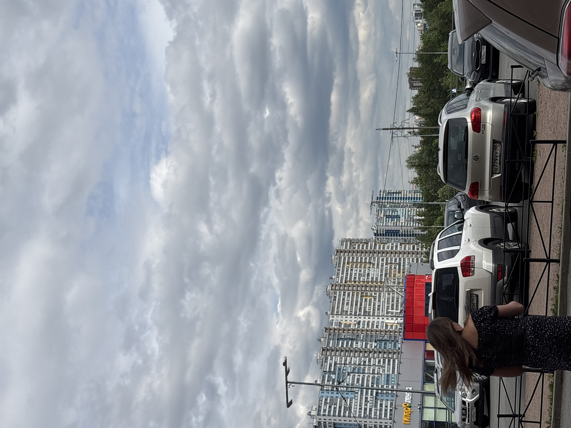 The image presents an urban street scene in Saint Petersburg, Russia, under a partly cloudy sky. Large gray and white clouds dominate the upper portion, with segments of lighter blue sky peeking through.
In the foreground, several cars are parked, separated from the viewer by a black metal fence. A woman with long, light brown hair, wearing a dark patterned dress, is partially visible on the right, standing behind the fence and looking towards the left.
Visible vehicles include a silver Volkswagen SUV with Russian license plate "H 779 KY 98" and "2.0 TSI" badging. Behind it, a dark Kia has the license plate "E 759 MO 198." Further left, a white Kia is parked, and a silver Nissan with license plate "A 400 HA 178" is seen closer to the bottom edge. The license plate regional codes (98, 198, 178) all correspond to Saint Petersburg.
Behind the parked cars, a line of trees runs across the mid-ground. In the background, numerous modern, multi-story apartment buildings with repeating window patterns form a dense urban landscape. A prominent street light or utility pole stands between the viewer and the buildings. A bright red rectangular structure is visible among the buildings.
Part of a commercial sign, reading "ГиперЛенд" (HyperLend), is visible in the lower-left section of the image. The scene depicts a daytime setting with diffused light due to the overcast conditions.
