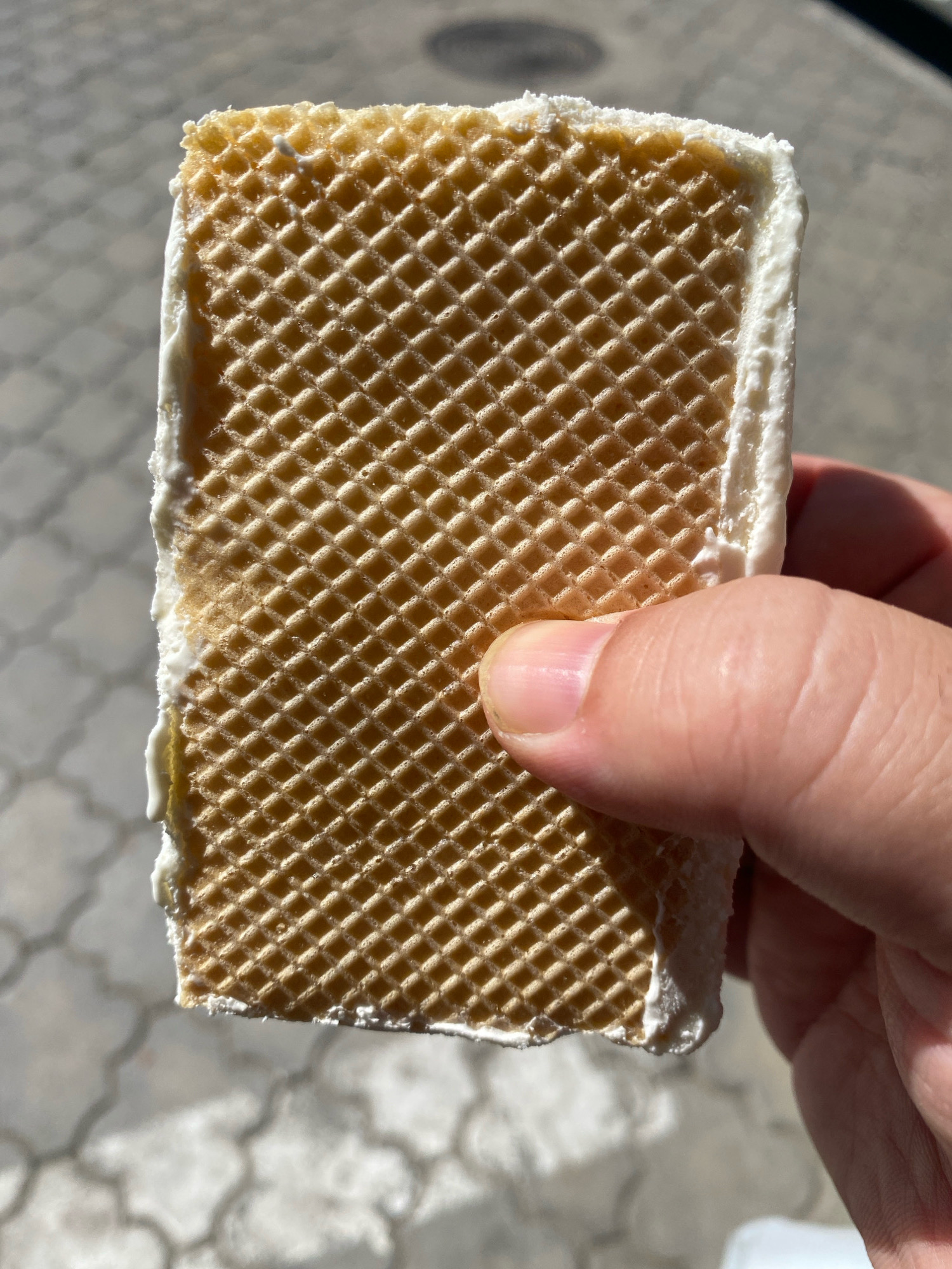 A close-up shot focuses on a rectangular wafer cookie, likely a part of an ice cream sandwich, held by a human hand. The cookie has a distinctive grid pattern. White, creamy filling is visible along the edges of the cookie, suggesting it is ice cream. The hand, with visible fingernails and skin texture, is gripping the cookie from the right side. The background is blurred and appears to be an outdoor setting with paved ground that has a repeating pattern, possibly a sidewalk or patio. Sunlight casts shadows, indicating it is daytime. The exact location in Chișinău, Moldova, cannot be determined from the visual information provided.