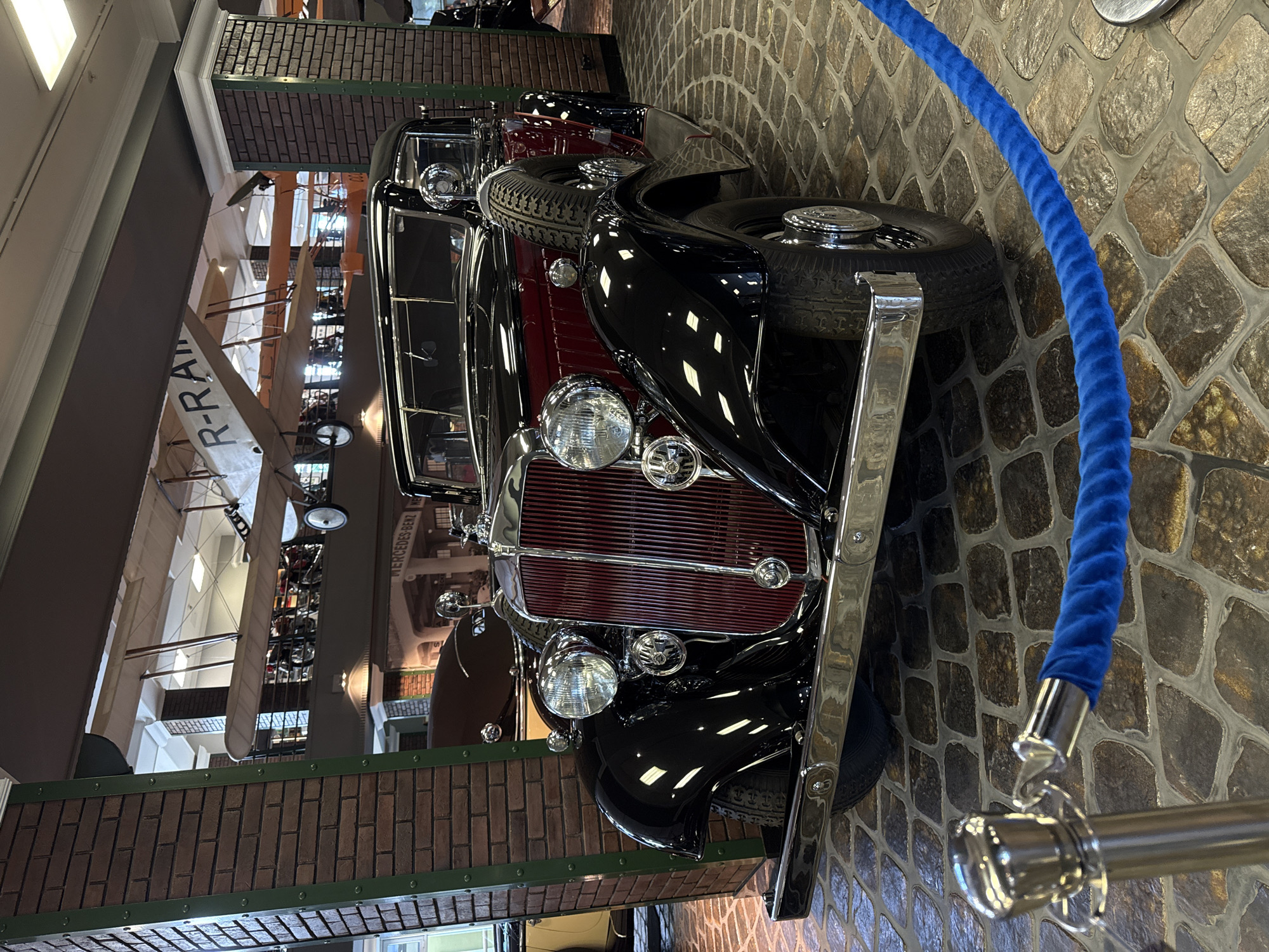 The image captures a vintage black and red convertible automobile displayed indoors on a cobblestone floor. The car features a prominent chrome grille, dual headlights, and a spare tire mounted on the side. In the background, several biplanes are suspended from the ceiling, suggesting a museum or exhibition setting. A brick pillar is visible on the left, and another on the right supporting an archway with a "MERCEDES-BENZ" sign. A blue velvet rope on a stanchion guides viewers. The scene appears to be within the Arkhangelskoye Estate Museum-Reserve in Arkhangel'skoye, Russia, known for its collection of historical vehicles and aircraft. There are no people visible in the frame.