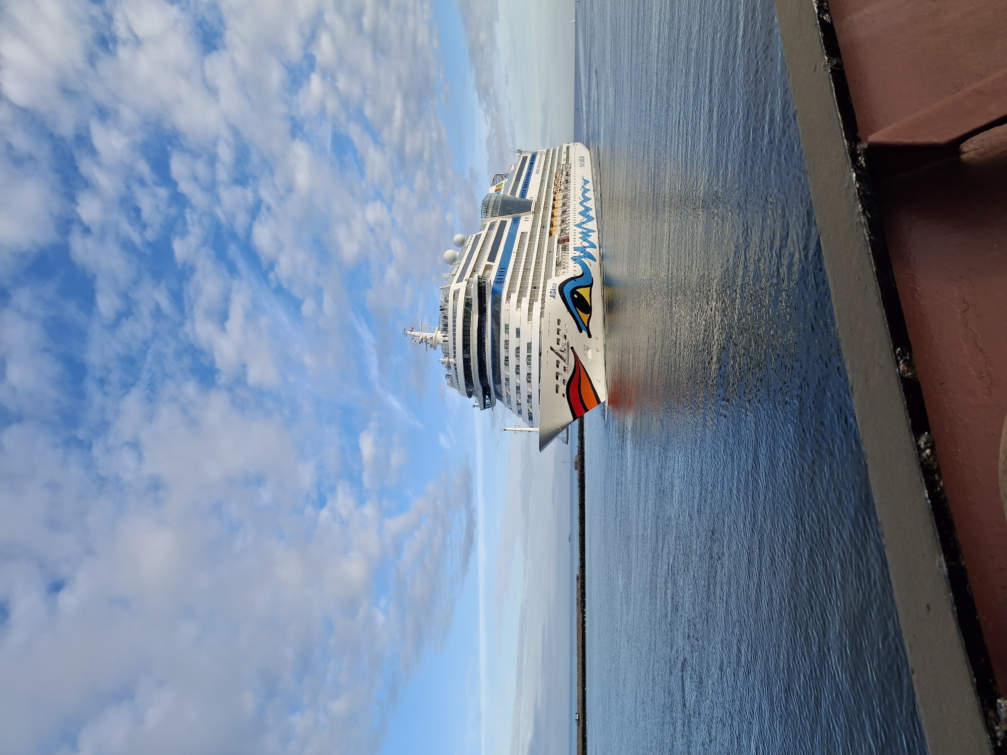 A medium shot depicts a large AIDA cruise ship in Rostock, Germany, on the Warnow River near Warnemünde. The ship is white with blue, yellow, orange, and red face art on the bow, and numerous rows of windows spanning multiple decks. The sea is calm and dark blue with a slight reflection of the ship. The sky is a light blue with scattered white clouds. In the foreground, a portion of a maroon structure, possibly part of the pier or another ship, is visible on the right. In the distance, a land mass can be seen through the horizon.