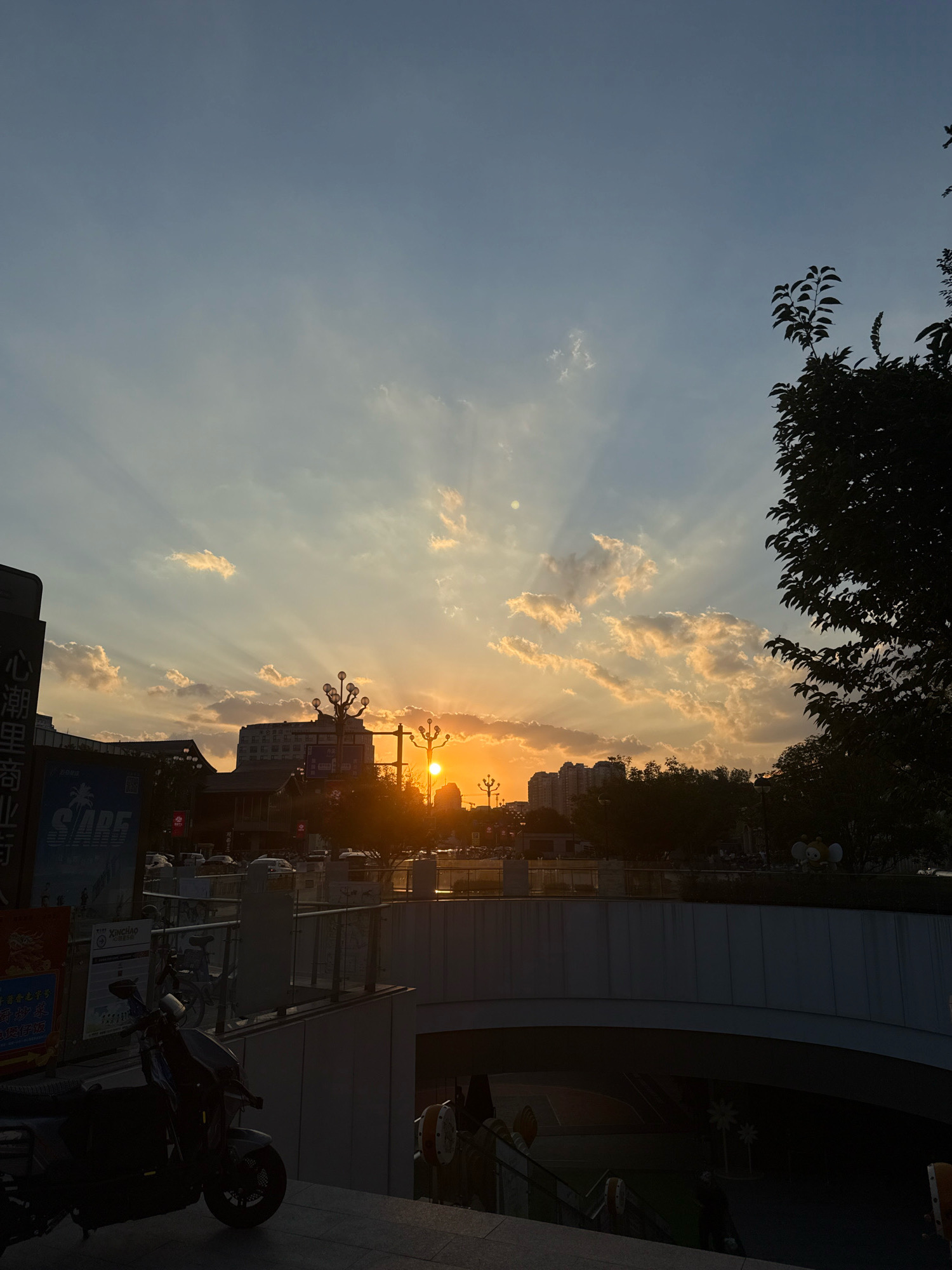 A sunset scene is captured in Yiwu, China, with the sun casting dramatic rays through scattered clouds. In the foreground, a dark, empty staircase leads down into a dimly lit underpass. A partially visible motorcycle is parked on the right side of the staircase. Above the underpass, a bridge or walkway is visible, lined with a metal railing. In the midground, silhouetted buildings and trees create a cityscape against the colorful sky. Several streetlights with globe-shaped lamps are prominent. The sky transitions from a pale blue at the top to warm orange and yellow hues near the horizon, where the sun is setting. The overall impression is one of a quiet evening in an urban environment.
