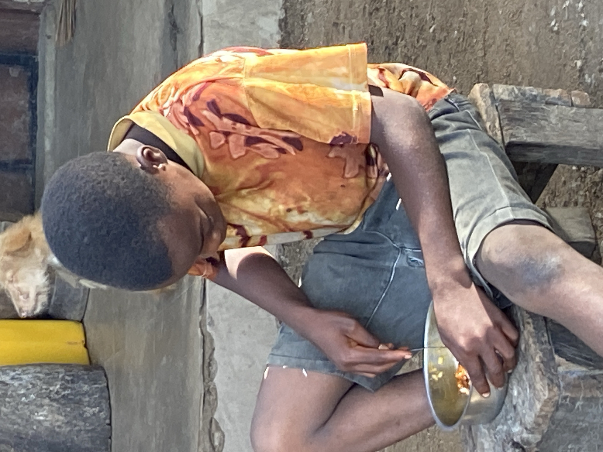 This image, taken in Mararaba, Nigeria, captures a young Black person in a casual outdoor setting during what appears to be bright daylight. The subject, likely a boy, is seated on a simple, rough-hewn wooden bench or stool, leaning forward with his head tilted down towards a metal bowl. He is wearing a vibrant orange, yellow, and brown patterned short-sleeved shirt and dark grey shorts.
The central activity shows him eating a meal from the bowl, using a small utensil (possibly a fork or spoon) held in his right hand. The bowl contains what appears to be a mixture of rice or grains with other small pieces of food. His focus is entirely on his meal.
In the lower-left corner of the frame, a light-colored animal, possibly a cat or a small dog, is visible, resting near the boy's left side. The background consists of rustic, light-colored walls or structures, suggesting a simple, possibly rural or semi-urban environment. A yellow object is also partially visible near the animal. The lighting indicates a sunny day. There is no visible text in the image.