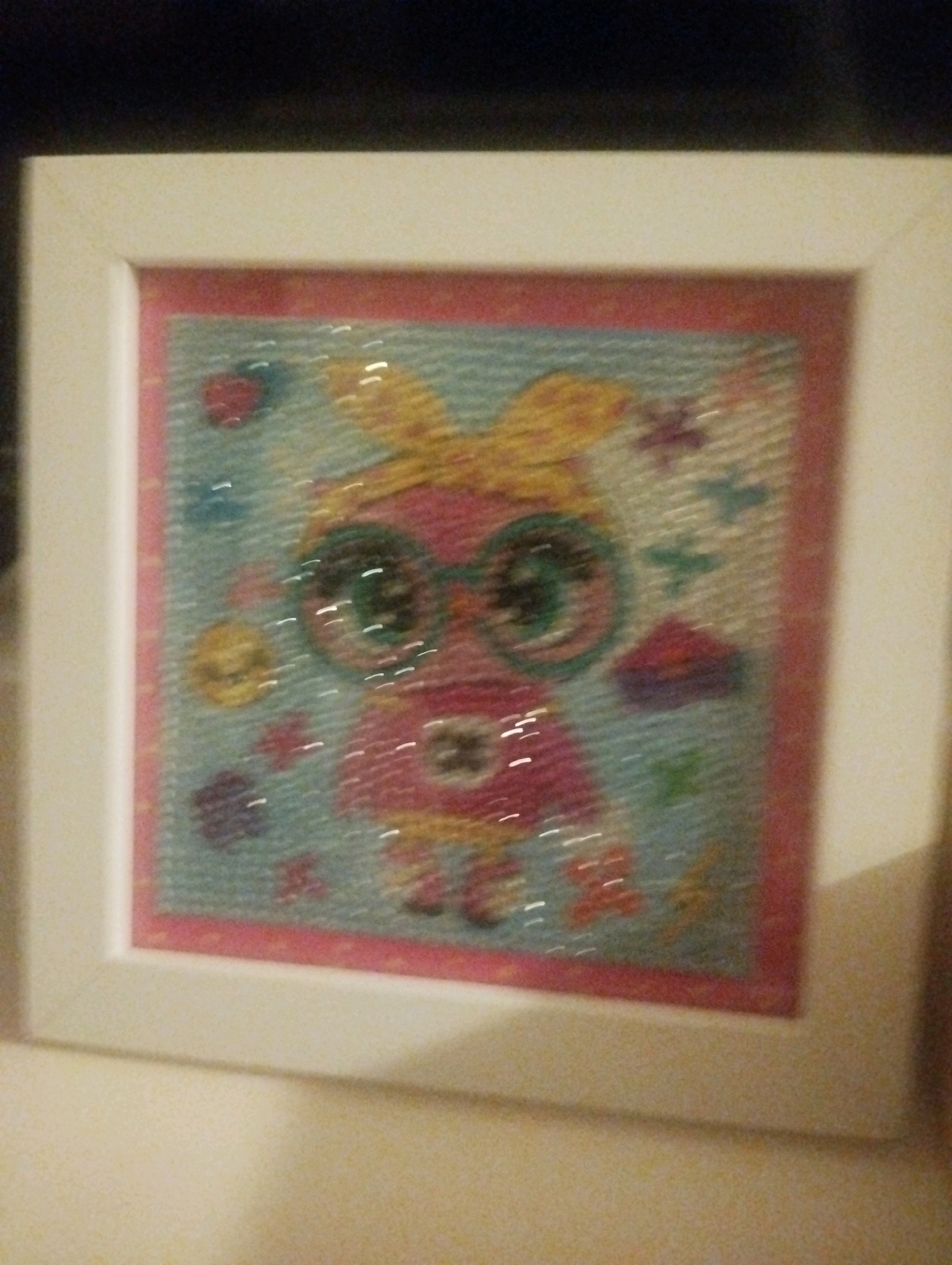 A framed diamond painting features a cartoon owl with large, sparkling green eyes and a yellow bow. The owl is pink with a white collar and a yellow shirt. It wears green-rimmed glasses. The background is light blue with scattered purple and red flowers and yellow circles. The frame is white and rectangular, and the painting is square. The image appears to be taken indoors in a residential setting in Ashdod, Israel, on a table.