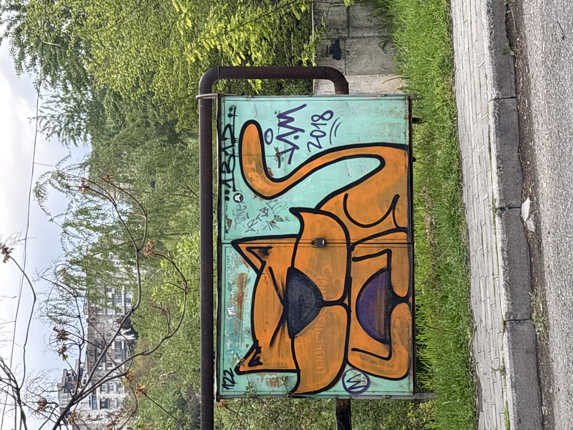 An outdoor shot captures a light teal-colored utility or electrical box adorned with vibrant graffiti art. The main subject of the graffiti is a large, cartoonish, orange cat-like creature with bold black outlines. The creature is depicted with one eye closed in what appears to be a wink, and purple accents are visible within its features, including an interior ear and another feature near its mouth. A long, curving tail extends upwards from the cat's body on the right side of the box.
Above the cat's tail, purple text reads "V.V.M" and "2018", indicating the artist's tag and the year of creation. On the bottom left corner of the box, the number "122" is visible. A small "W" in a circle is also present near the bottom right of the cat's face. The metal box itself shows signs of weathering, with some rust and faded paint beneath the artwork. A rusty metal pipe runs horizontally across the top of the box and vertically along its left side.
The box is situated next to a paved sidewalk made of rectangular blocks, which borders an asphalt road on the right. Lush green grass grows between the box and the sidewalk. In the background, abundant green foliage from trees and bushes creates a vibrant backdrop. Through the trees, a tall, multi-story building with a distinctive architectural style can be seen in the distance, suggesting an urban setting, consistent with Tbilisi, Georgia. The sky above is partly cloudy, and the scene is illuminated by daylight, indicating a daytime shot. No human activity is depicted.
