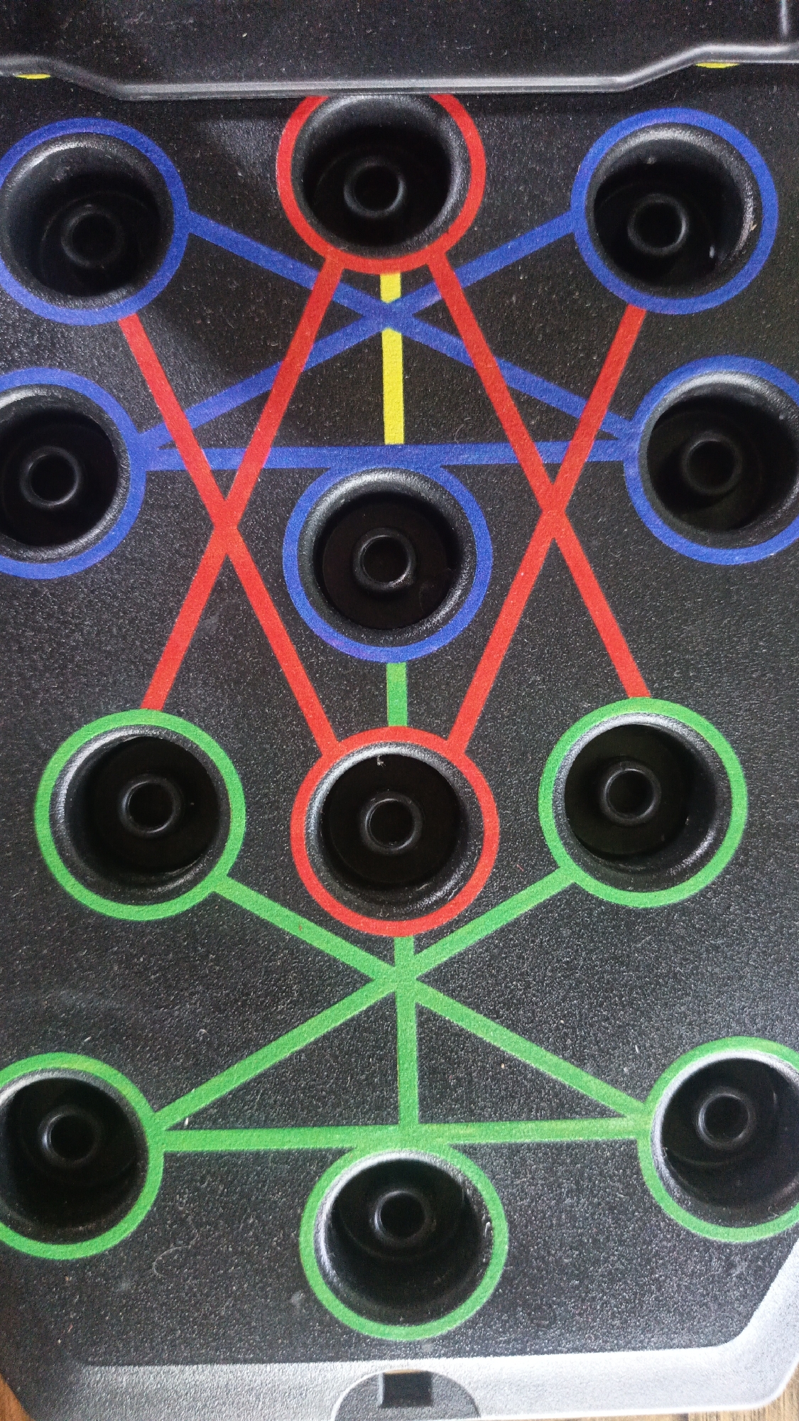 The image is a close-up, top-down view of a black push-up board. The board is marked with a geometric pattern of colored lines: red, blue, and green. These lines connect multiple circular indentations, which appear to be holes or grips for hands. There is also a small section of yellow line visible. The surface of the board has a textured finish. The provided location, Sungai Rasau, Indonesia, is irrelevant to the visual content of the image. There are no people, actions, or interactions depicted. The scene is entirely an inanimate object.