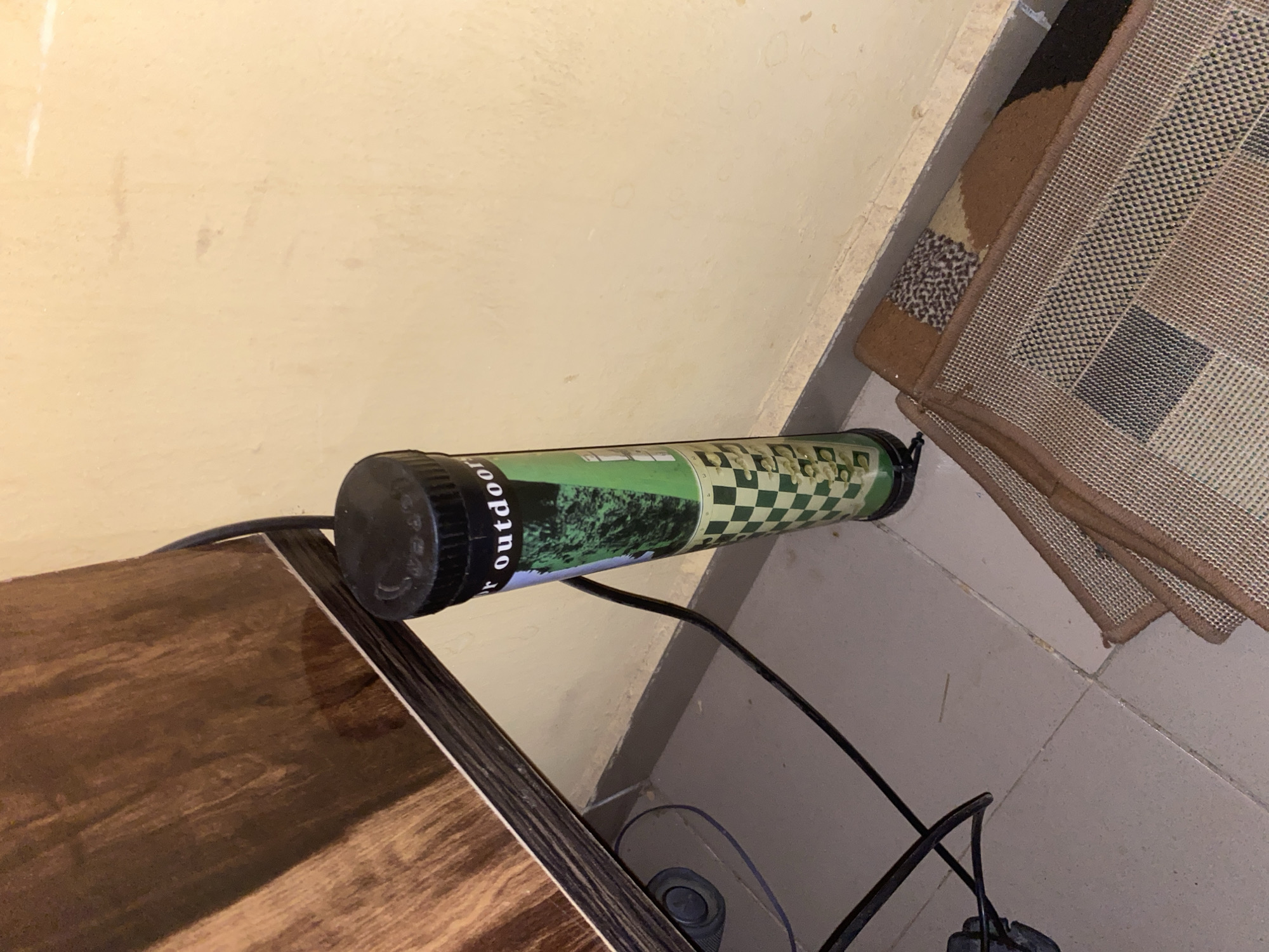 The image shows an "Outdoor Router" device positioned on top of a wooden cabinet, near a wall and a rug. The environment appears to be an indoor corner, possibly in a room within a building. The router is cylindrical with a black cap and has green and checkered patterns on its body. A black power cable is connected to it, leading away from the device, and another cable is visible on the floor. The rug next to the wall features a geometric pattern. The wall is plain and painted in a light beige color. The floor is tiled with light-colored tiles. The specific location within Hanwa, Nigeria, is not discernable from the image.