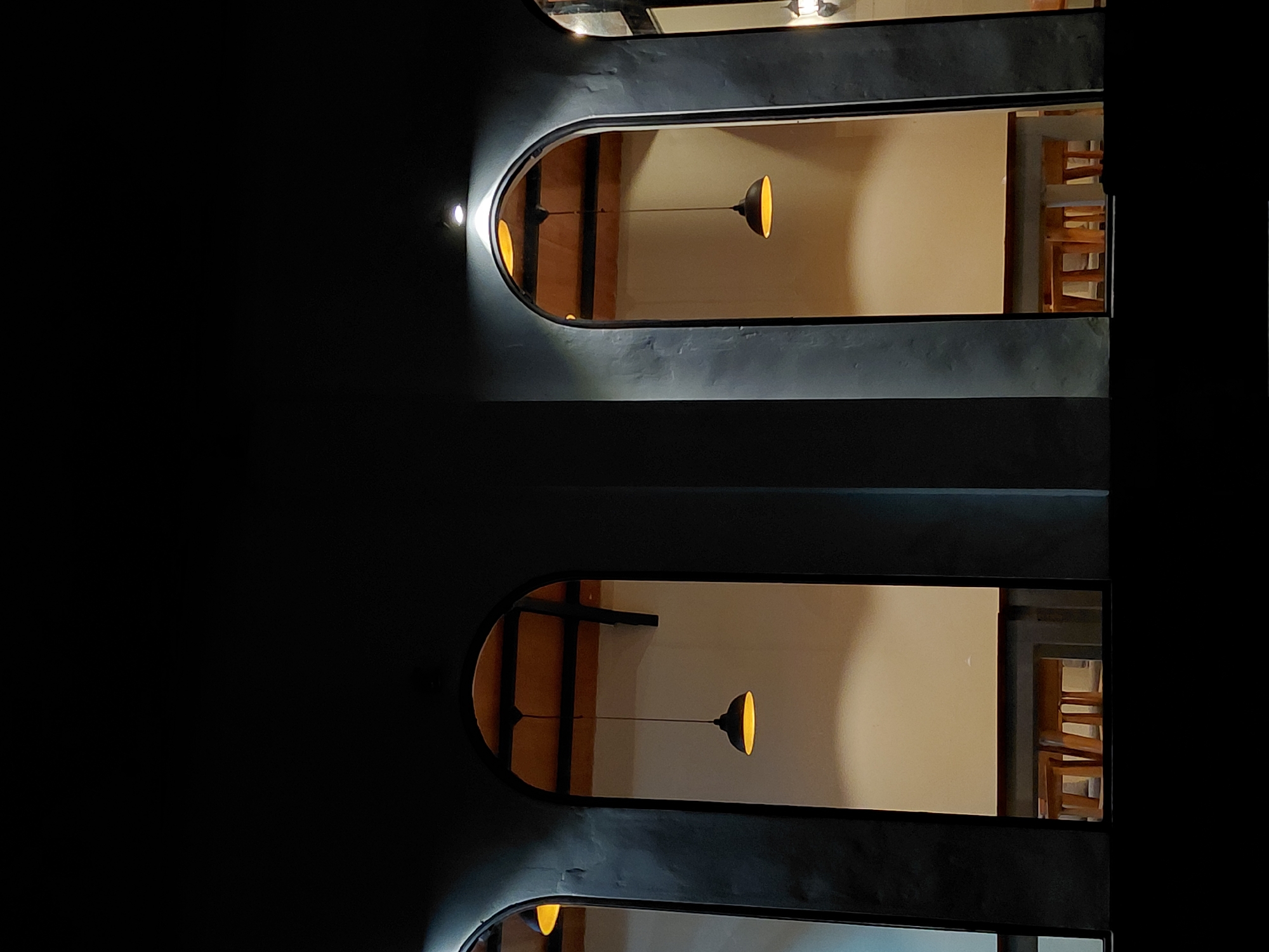 The media shows an interior view of a dimly lit space, possibly a restaurant or cafe in Kintamani, Indonesia, at night. Two horizontal, elongated windows with rounded ends are visible. Each window looks into a separate, similar room.
The upper window reveals a room with a light-colored wall and wooden beams or furniture. A single pendant light fixture with a black shade and a yellow-lit interior hangs in the center of the visible space. To the right, several wooden chairs are stacked. A small, bright light source is visible on the wall to the left of the window, casting a strong spotlight.
The lower window shows a similar scene. A pendant light fixture, identical to the one in the upper window, hangs in the center. The wall behind it is also light-colored. To the right, stacked wooden chairs are visible, similar to those in the upper section. A small, bright light source is also present on the wall to the left of this window.
The overall impression is one of modern, minimalist interior design with a focus on warm, ambient lighting. The darkness surrounding the windows suggests it is nighttime, and the absence of visible people indicates the space is either closed or empty at the time of the photograph. The location context of Kintamani, Indonesia, adds a cultural layer, though no specific Indonesian cultural elements are explicitly visible in the immediate frame.