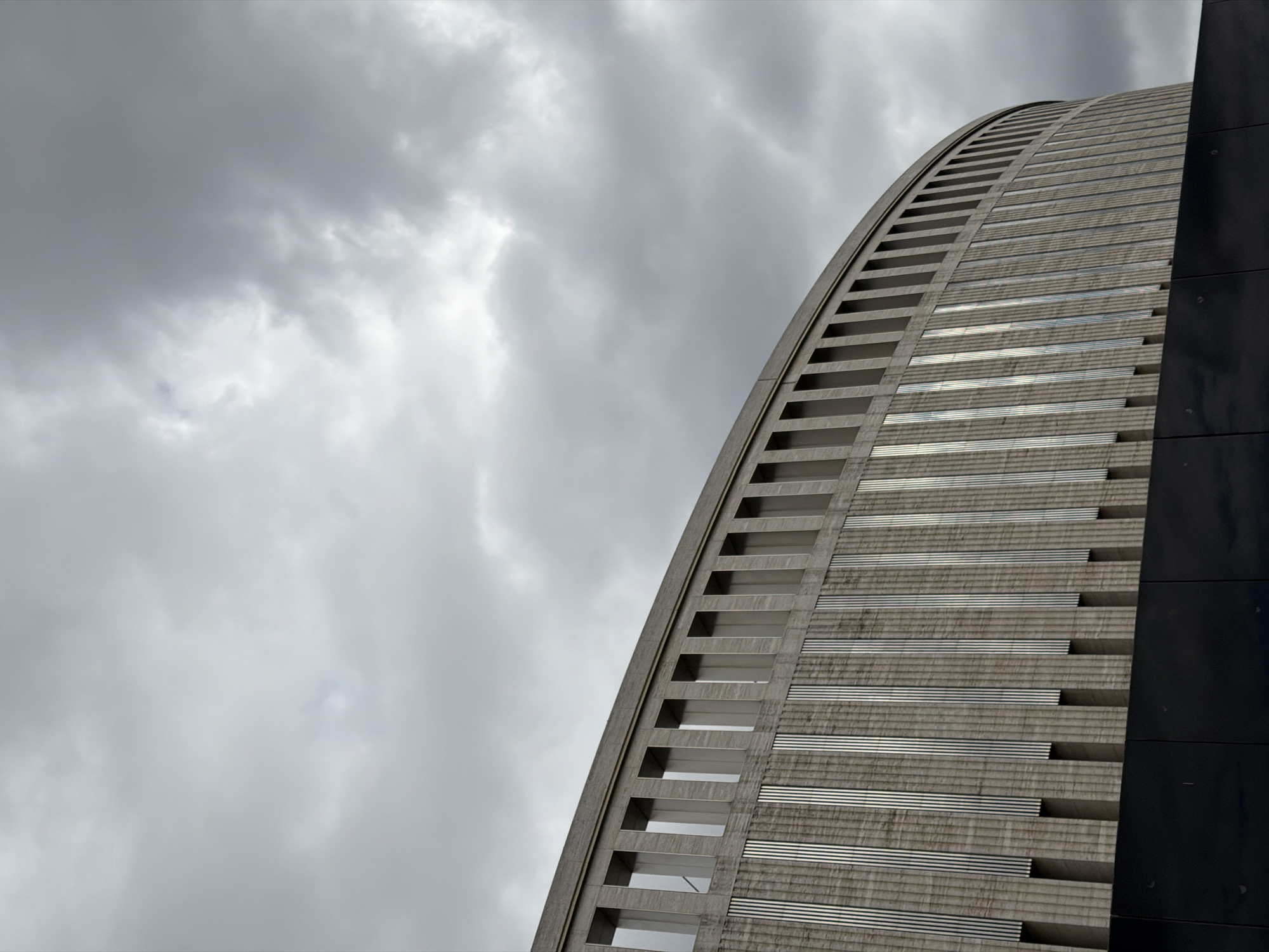 The image presents a low-angle view of a curved, high-rise building in Krasnodar, Russia. The building's facade is segmented with horizontal bands and rectangular openings, suggesting a modernist architectural style. A dark, smooth surface adjoins the curved portion of the building on the right side of the frame. The background is dominated by a cloudy sky, hinting at overcast weather conditions. The overall tone of the image is muted due to the prevalent gray hues.
